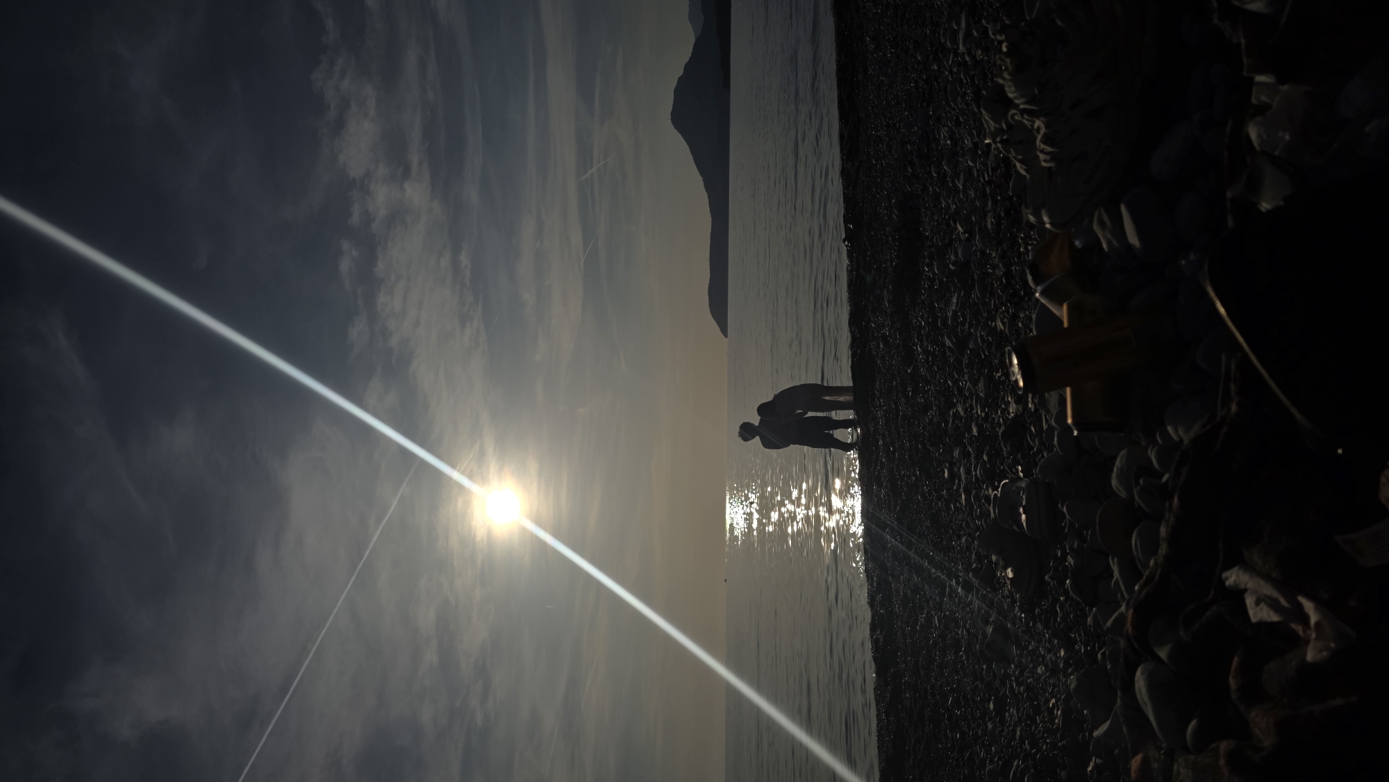 Two people stand on the rocky beach of Žukotrlica in Bar, Montenegro. The sun shines brightly, casting a strong glare on the water. The silhouettes of the individuals are visible against the sunlight. A mountain rises in the background. The sky is streaked with clouds and contrails.