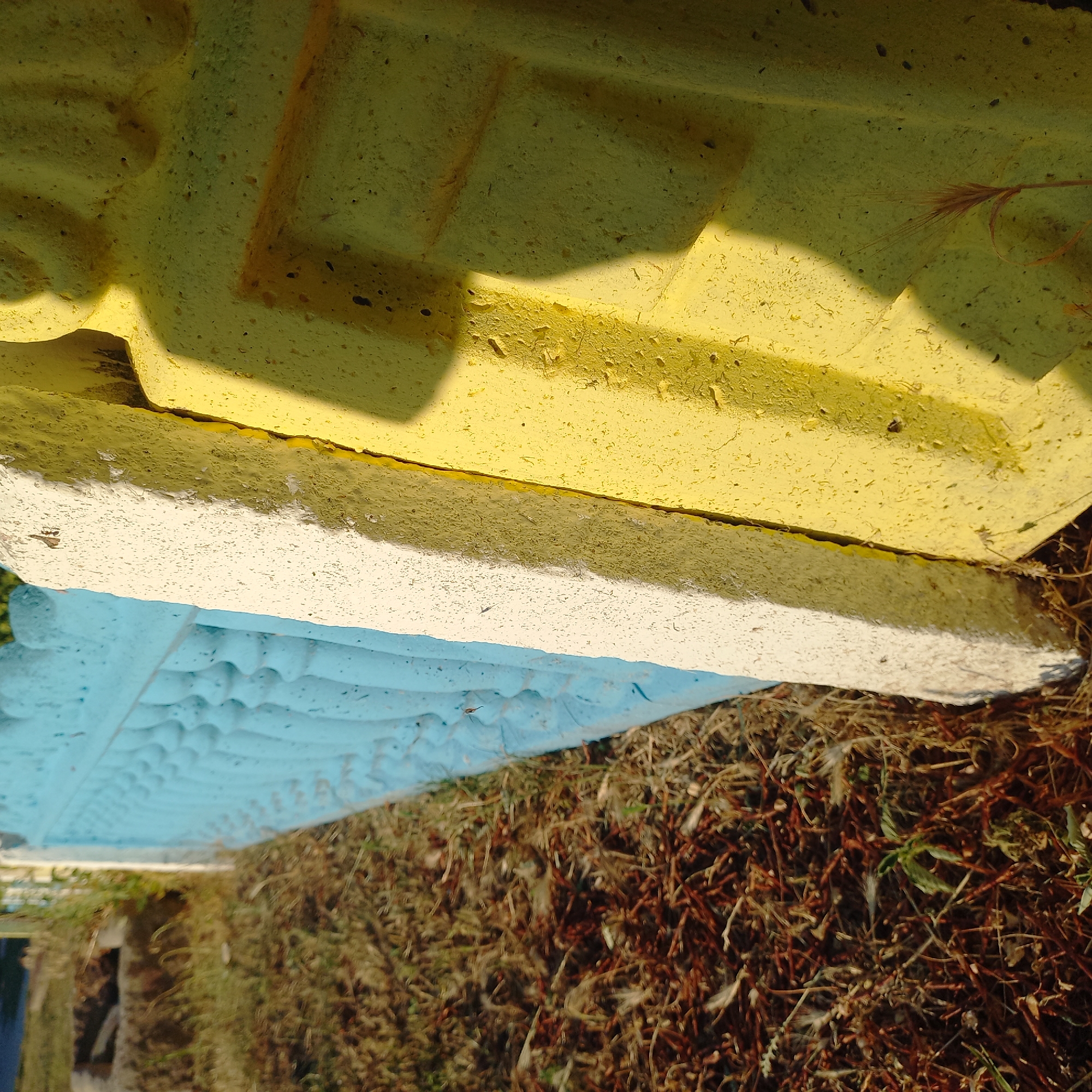 The image shows a section of a decorative fence in the city of Klyntsi, Ukraine. The fence is constructed from concrete elements painted in alternating colors of light blue, white, and yellow. The blue section on the left features a repeating pattern of ornate balusters. Adjacent to it, a white rectangular concrete element is visible, and to the right, a yellow concrete element with a geometric pattern is depicted. The fence is situated outdoors, with dry, brown grass and some sparse green vegetation covering the ground in the foreground. The lighting suggests it is daytime, with shadows cast by the concrete elements. There are no people or actions depicted in the image.