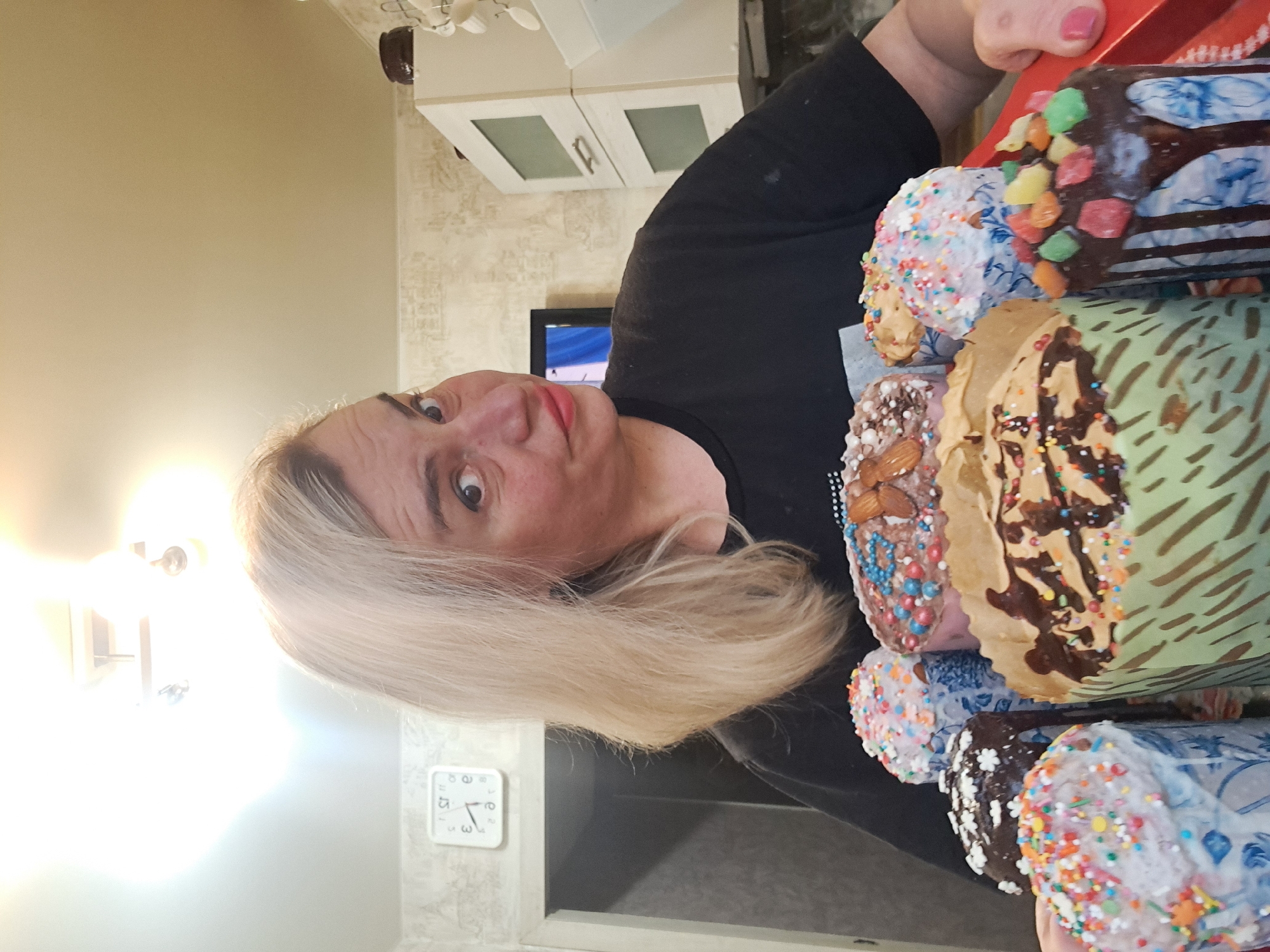 A woman with fair hair is holding a stack of decorated Easter cakes (kulichi) in what appears to be an indoor setting, likely a kitchen. The cakes are decorated with various toppings including icing, sprinkles, nuts, and candied fruits. The woman is wearing a black t-shirt and looking at the camera with a slight smile. In the background, there are kitchen cabinets, a television, a clock on the wall, and a light fixture. Based on the available information, it's not possible to conclusively determine if the location is in the city of Elektrostal, Russia.
