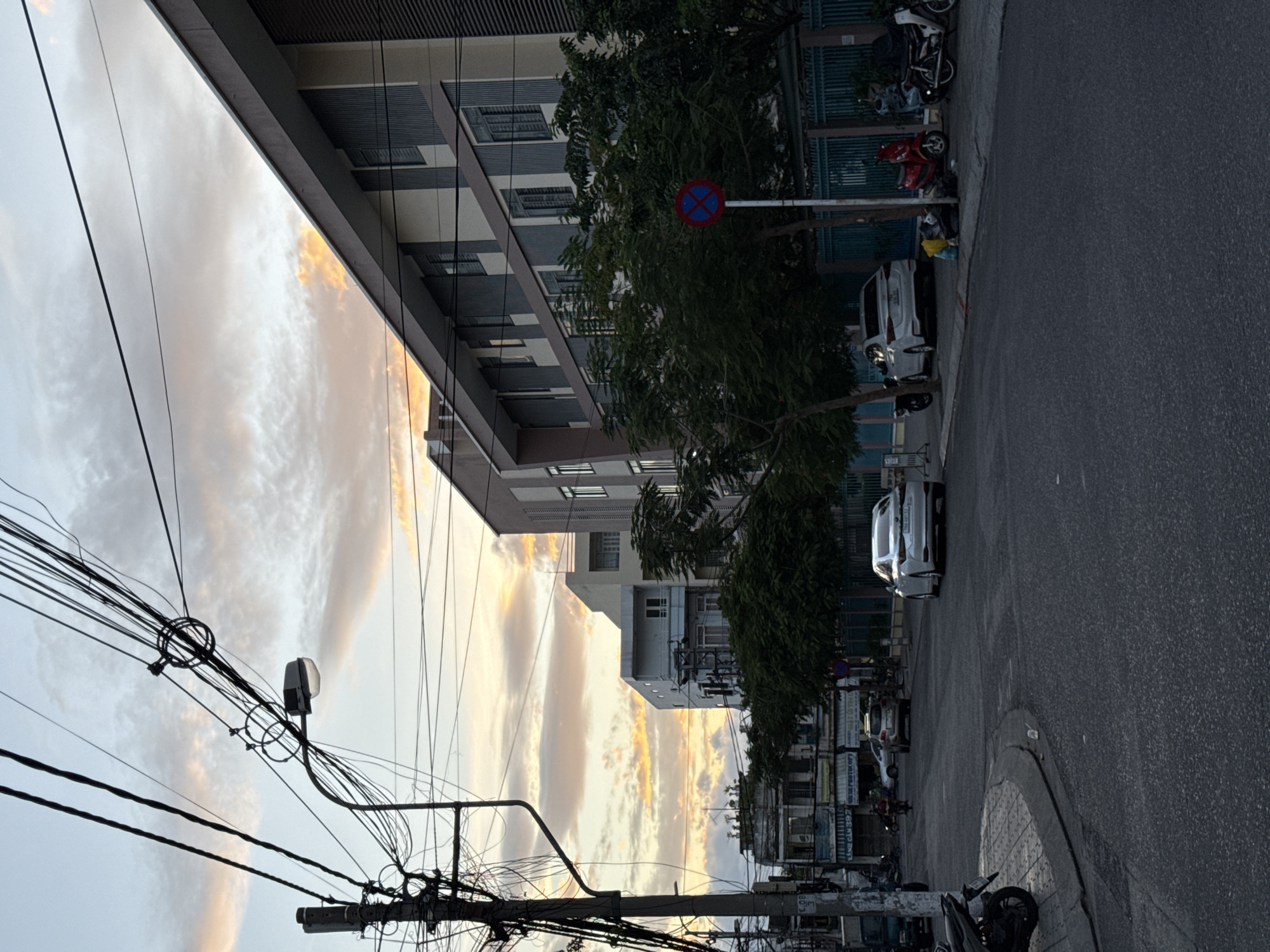 The image depicts a street scene in Nha Trang, Vietnam, at dusk. A modern building with horizontal grey and beige siding dominates the right side of the frame, its angled roofline cutting across the sky. Across the street, older, multi-story buildings are visible, with a power pole heavily laden with tangled wires in the foreground on the left. The sky is filled with scattered clouds tinged with orange and pink from the setting sun. Several vehicles, including cars and motorbikes, are parked along the street and on the pavement. One white sedan is parked in front of the modern building, while a red scooter and another motorbike are visible to its right. In the foreground, the asphalt road curves to the left, with a section of paved sidewalk. A circular traffic sign with a red border and blue center is posted on a pole near the modern building. The overall impression is of a typical urban streetscape transitioning from day to night.
