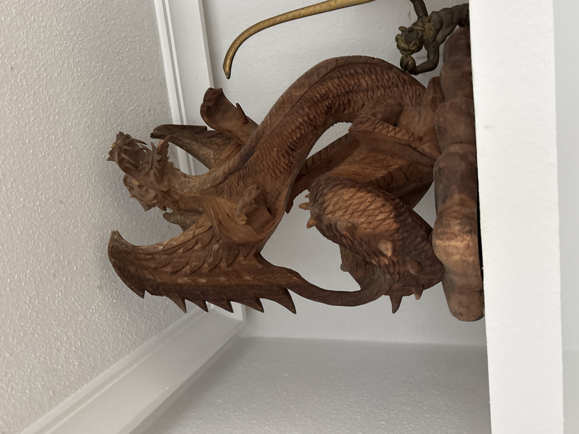 A wooden sculpture of a dragon is displayed on a white shelf, with a white textured ceiling above. The dragon is carved with detailed wings, scales, and an open mouth with visible teeth. To the right of the dragon, there is a smaller, dark bronze figurine of a creature with a human-like torso and legs, holding a spherical object. A thin, curved golden rod-like object is also partially visible to the right of the dragon, extending upwards. The scene appears to be an indoor setting. The location is not specified to be in the city of Orihuela, Spain.
