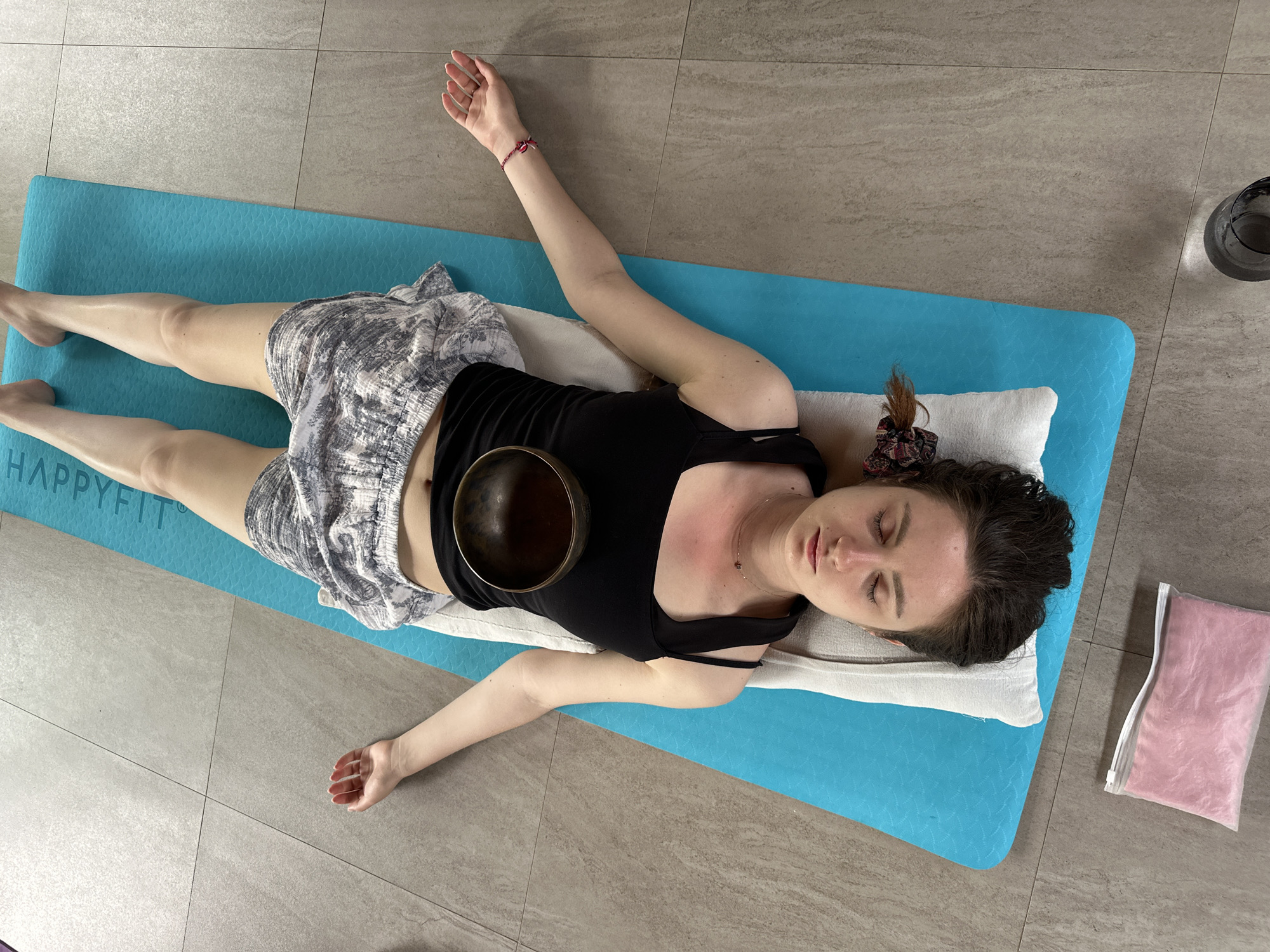 A woman is lying in the Shavasana pose on a blue yoga mat on a tiled floor. She is wearing a black tank top and patterned shorts. A singing bowl rests on her stomach. Her head is on a white pillow. A clear bag of pink material is on the floor next to the mat. A partially visible glass vase is in the background. The word "HAPPYFIT" is visible on the mat. This image was taken in Sukawati, Indonesia.