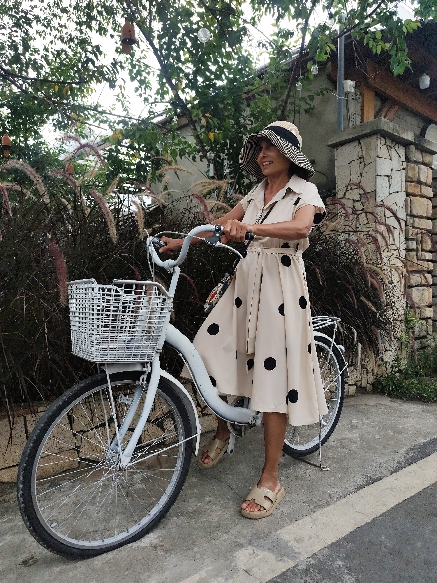 A woman stands next to a white bicycle with a white basket in front. She is wearing a light-colored, polka-dotted dress and a wide-brimmed hat. The bicycle has a kickstand supporting it. The background features lush greenery, a stone wall, and a building with a wooden roof. The scene appears to be outdoors, possibly in a garden or courtyard. The woman is looking to her left and smiling, suggesting a relaxed and pleasant mood. The lighting suggests it is daytime, and the presence of greenery and the stone architecture might indicate a location in Da Lat, Vietnam. The woman's attire and the bicycle suggest a casual outing.