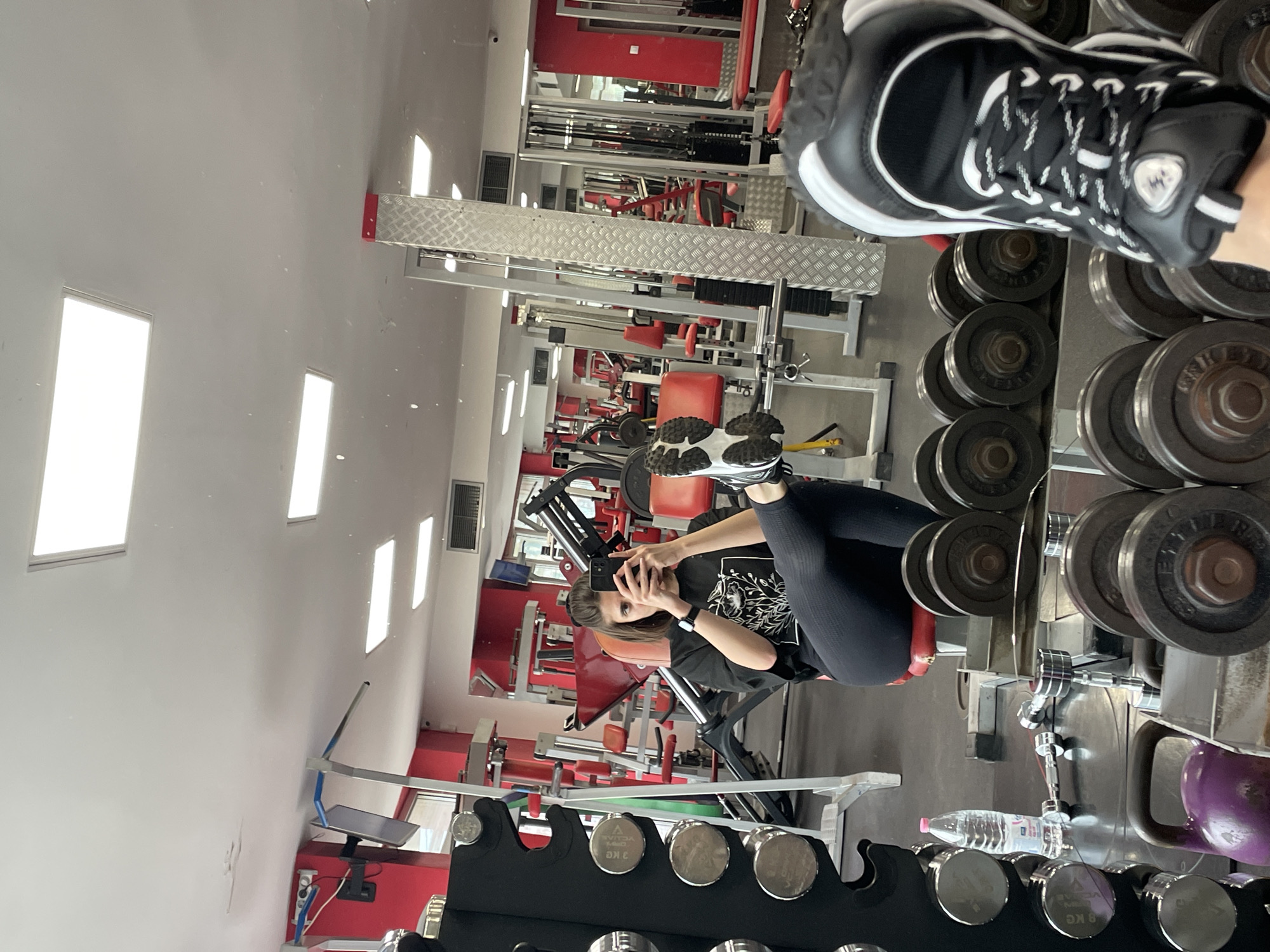 A person in black leggings and a black t-shirt is taking a selfie in a gym. The gym is filled with various exercise machines and weights. The image is taken in a mirror, with dumbbells in the foreground. A water bottle sits below the dumbbells. The gym setting suggests this is a commercial establishment. The location is Sofia, Bulgaria.