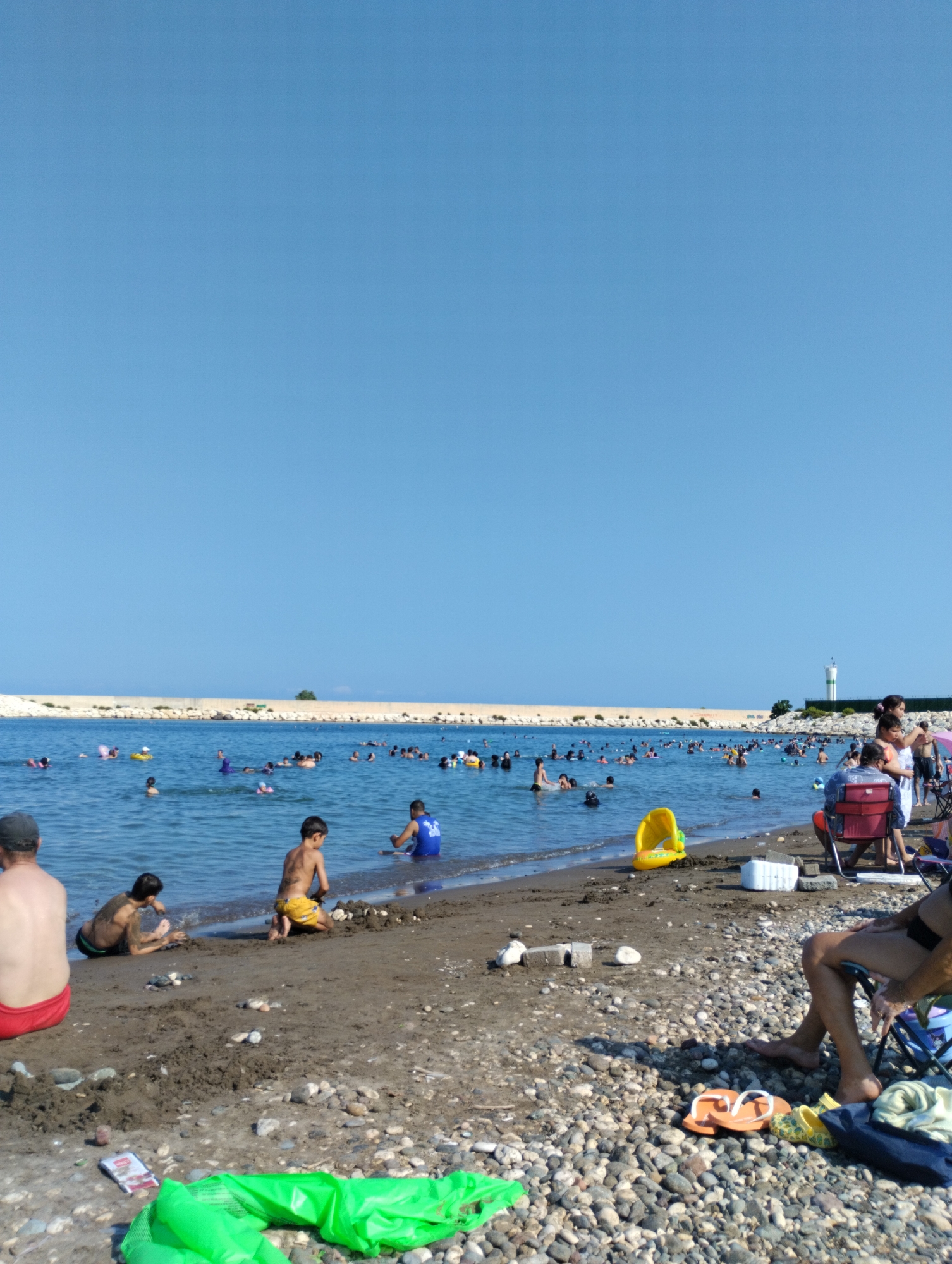 The image depicts a crowded beach scene on a sunny day. In the foreground, several people are relaxing on the pebbly shore. A man wearing red shorts is partially visible on the left. Two young boys are playing in the wet sand near the water's edge, one in a yellow swimsuit and the other in a green swimsuit. To the right, a person is seated in a red chair, and another individual, wearing a dark swimsuit, is sitting on a beach chair, with their legs crossed and resting on the pebbles. Flip-flops and a green inflatable object are scattered on the ground. The middle ground is dominated by the blue sea, filled with numerous people swimming and wading. Some individuals are using inflatable rings. The background shows a long, light-colored breakwater or sea wall extending into the sea, with a white lighthouse visible on the far right end of it. The sky is clear and bright blue. The location is identified as a beach in Erdemli, Turkey.