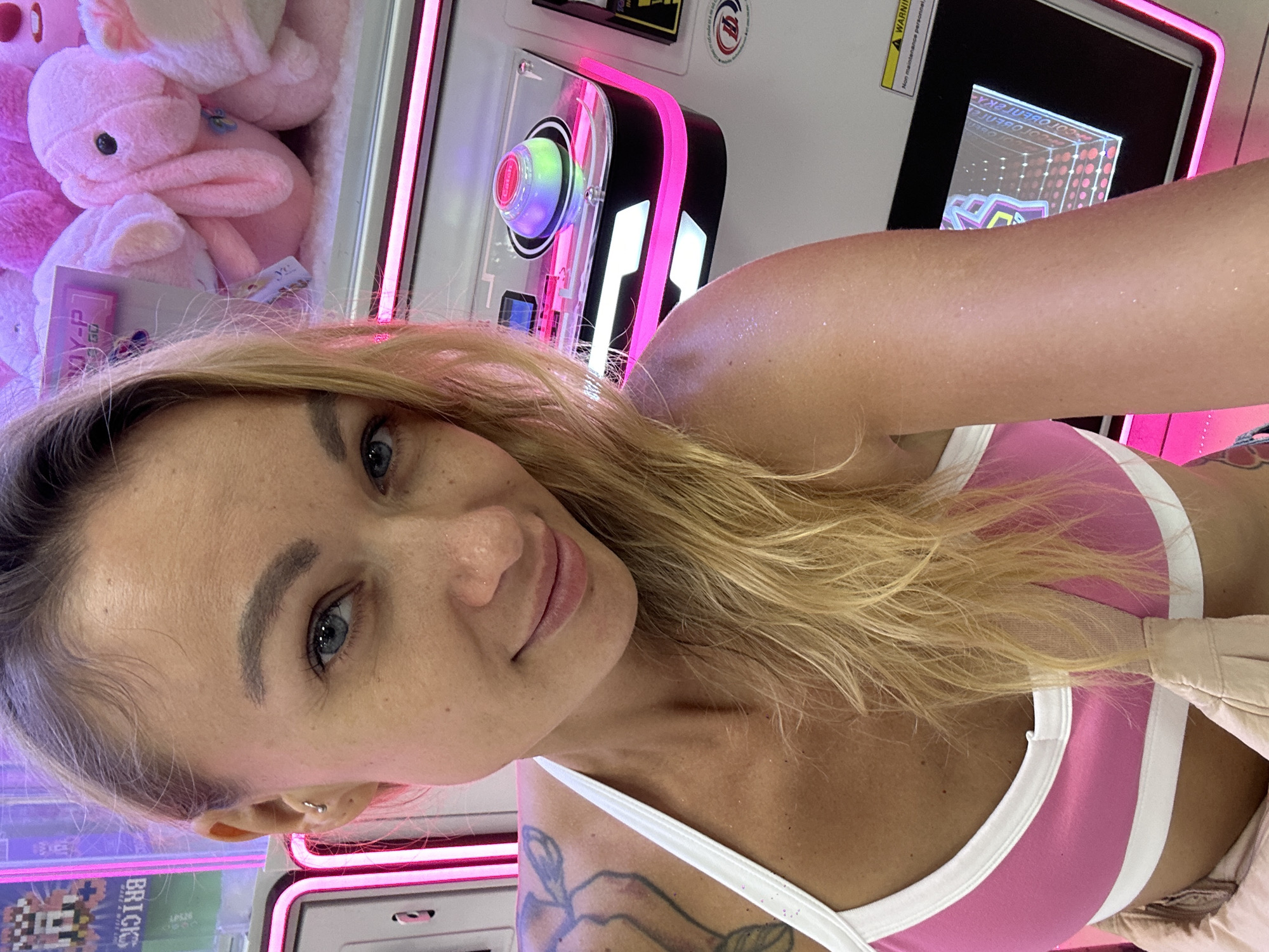 A young woman with blonde hair and blue eyes is taking a selfie in front of a claw machine in what appears to be an amusement center. She is wearing a pink and white sports bra. The claw machine is filled with pink stuffed rabbits and has brightly colored LED lights. The background shows other arcade games and signage, including a sign that says "BRICK". The lighting in the scene is predominantly pink, creating a vibrant and playful atmosphere.