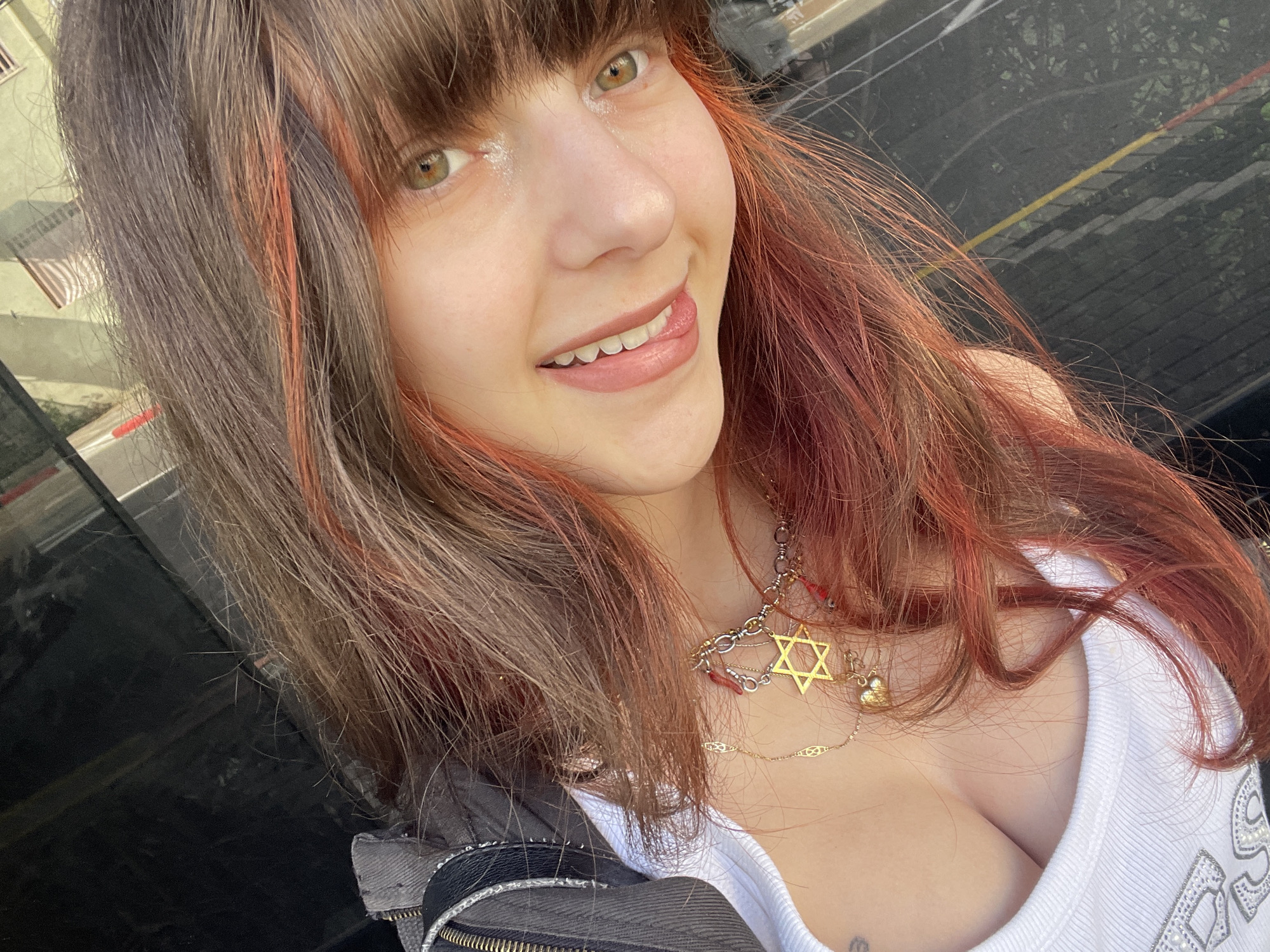 A young woman with brown hair and red highlights is smiling at the camera. She has bangs and hazel eyes. She is wearing a white tank top and a necklace with a Star of David pendant. A jacket is visible over her shoulders. The location appears to be an urban environment, potentially Petah Tikva, Israel, based on the presence of buildings and street markings in the background.