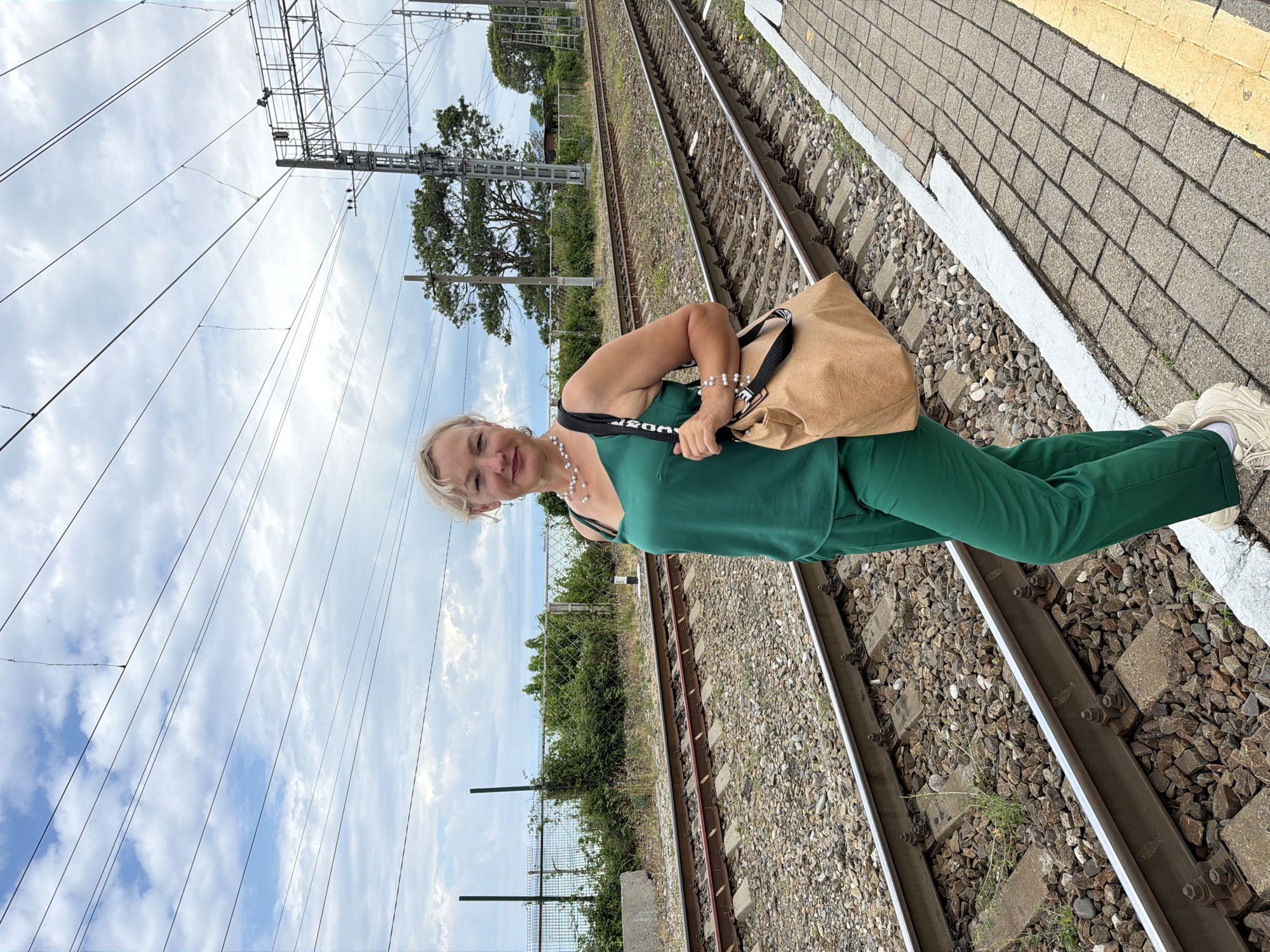 A blonde woman with light skin stands on a train platform in Shepsi, Russia. She is wearing a green tank top and matching pants, with white sneakers. She is carrying a large tan tote bag with a black strap. The woman is standing next to a set of train tracks, with lush green vegetation and trees in the background. The sky is partly cloudy.