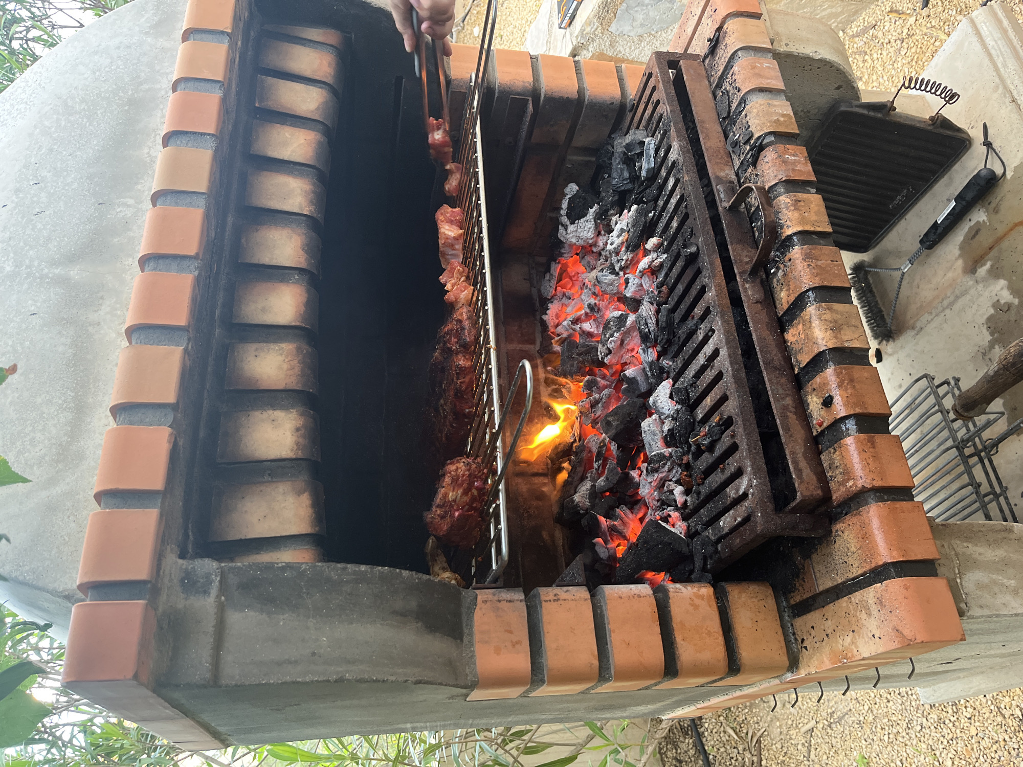 The image shows a brick barbecue in what appears to be a residential setting, likely in El Poble Nou de Benitatxell / Benitachell, Spain. The barbecue is constructed of red brick with a concrete base. There are two compartments, one with meat on a grill rack and the other with hot coals burning inside a metal grate. A hand holding tongs is visible, lifting or adjusting the meat. To the right of the barbecue are additional grill racks, a grill pan, and a grill brush, suggesting the barbecue is in use. The ground is covered with small stones and plants are visible. The weather conditions are not discernible from the image.