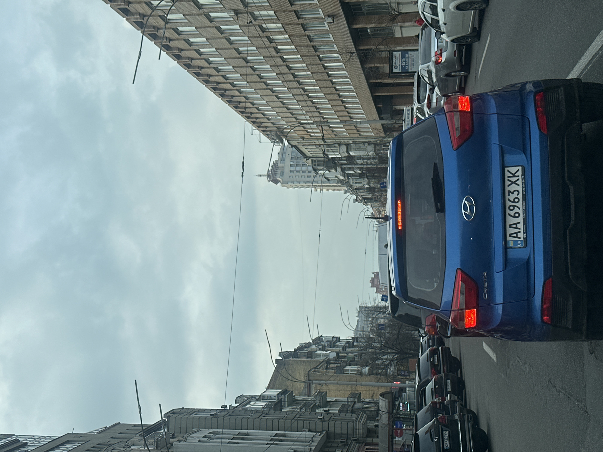 The image captures a street scene in Kyiv, Ukraine, with a focus on traffic congestion. A blue Hyundai Creta SUV with license plate "AA 6963 XK" is in the foreground. The vehicles are stopped in a line, with other cars and buses visible ahead. Several tall buildings line the street, with visible overhead power lines. The weather appears overcast and the sky is grey. The street includes commercial buildings, indicated by signs with text.