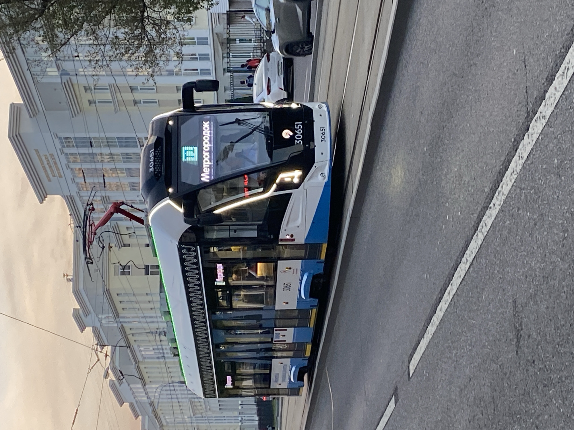The image shows a modern tram with the number 30651 and the destination "Метрогородок" (Metrogorodok) displayed on its front. The tram is on a city street with tram tracks. In the background, there are several multi-story buildings with a classical architectural style, suggesting an urban environment. There are also a few cars visible on the road and some pedestrians in the distance. The sky appears to be overcast, possibly at dusk or dawn, with soft, diffused light. The tram has a distinctive black, white, and blue color scheme with green accent lighting along its roof. The overall scene depicts a typical urban transit environment in Moscow, Russia, given the Cyrillic text on the tram and the architectural style of the buildings.