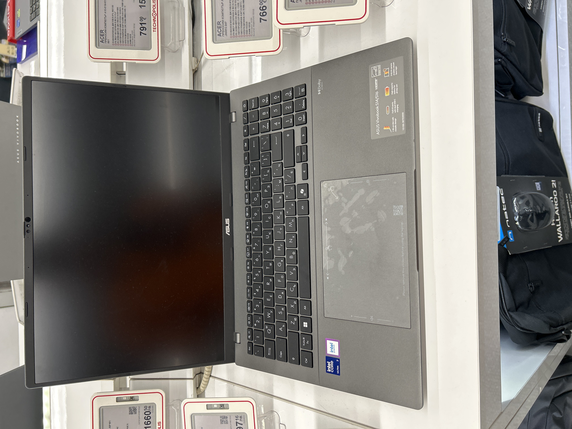 The image depicts a gray ASUS Vivobook laptop on display in a store, presumably in Sofia, Bulgaria, based on the Cyrillic script on the adjacent price tags. The laptop's screen is on, but the image displayed is dark. The keyboard shows Cyrillic characters alongside the Latin alphabet. Fingerprint smudges are visible on the touchpad. Stickers indicating "Intel Core Ultra 7" and "Dolby" are affixed to the chassis. Price tags for Acer laptops are visible nearby. Next to the laptop are black bags and a box for a "natec WALLAROO 21" product.