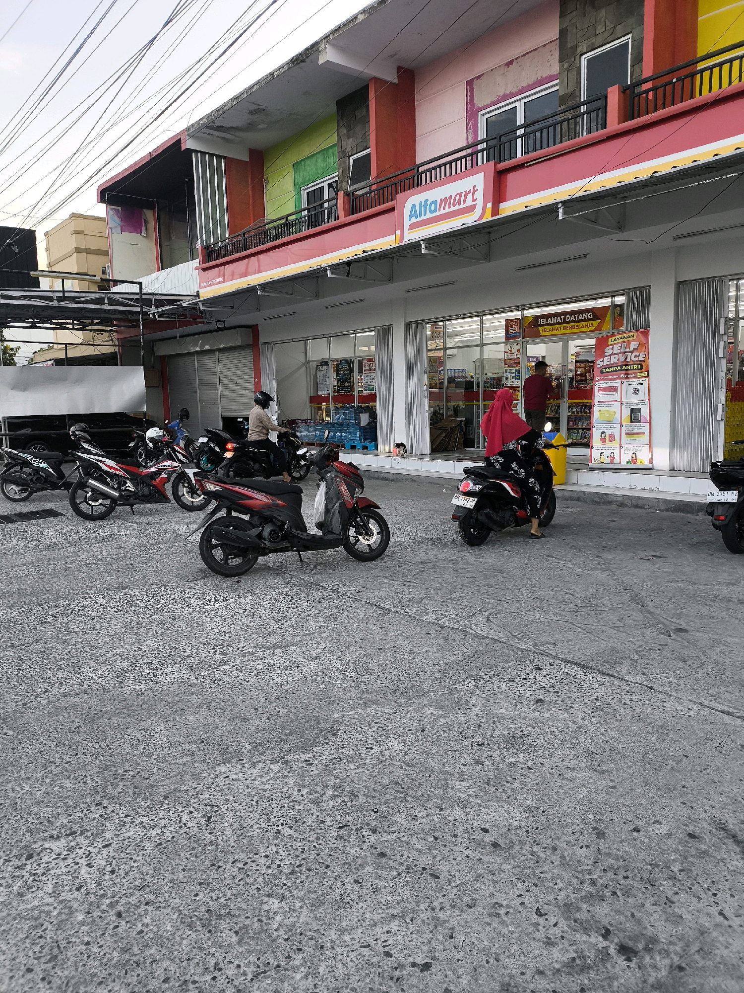 The image depicts the exterior of an Alfamart convenience store, likely located in the city of Gorontalo, Indonesia. The store features a red and yellow facade with prominent signage. Several motorcycles are parked in front of the store, indicating that customers have arrived by this mode of transportation. A few individuals are visible, including one person wearing a red hijab and another in a maroon shirt. One person is standing near the store entrance, and another is seated on a motorcycle. The ground in front of the store appears to be paved with textured concrete. The overall scene suggests a typical day at a neighborhood convenience store in an Indonesian city, with customers coming and going.