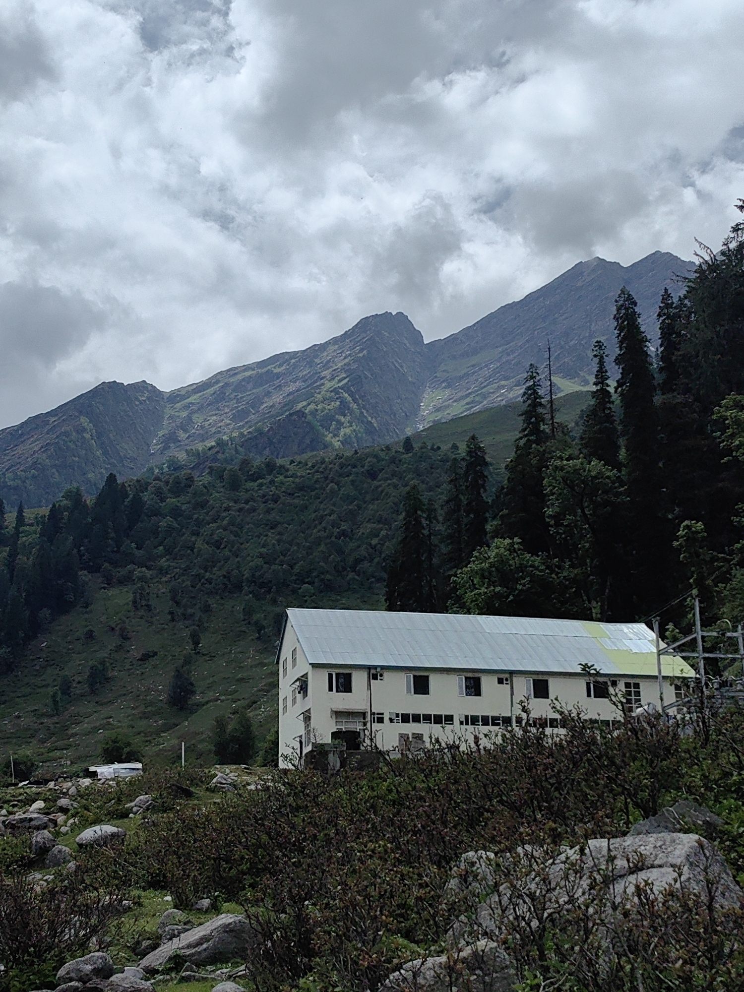 The image depicts a long, two-story building with a light-colored facade and a metal roof, situated in Dhundi, India. The building is surrounded by a natural, mountainous landscape characterized by steep, verdant slopes and a cloudy sky. In the foreground, there are rocks and dense, low-lying vegetation. In the background, imposing mountains rise, covered in lush greenery and interspersed with scattered trees. The scene is predominantly green and gray due to the foliage and the overcast weather. There are no visible people or animals in the image.