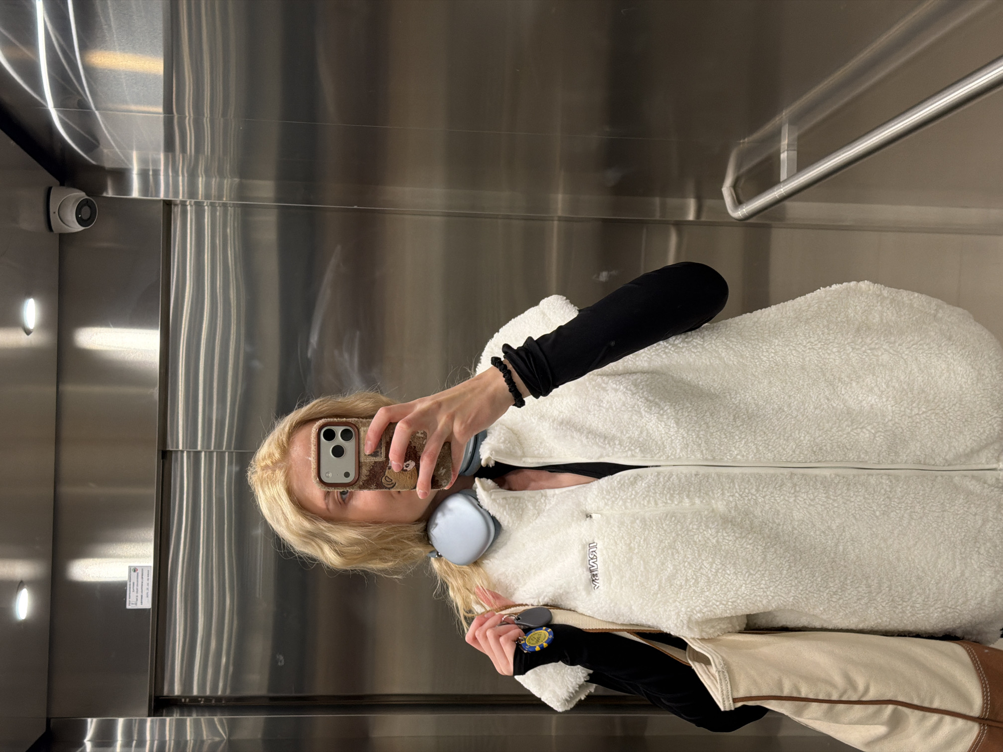 A woman is taking a selfie in a metal-walled elevator. She is wearing a white, fuzzy jacket and black sleeves, and has headphones around her neck. She holds a phone up to take the photo. Visible on the left wall of the elevator is a security camera. The location of this photo is not discernible from the image.