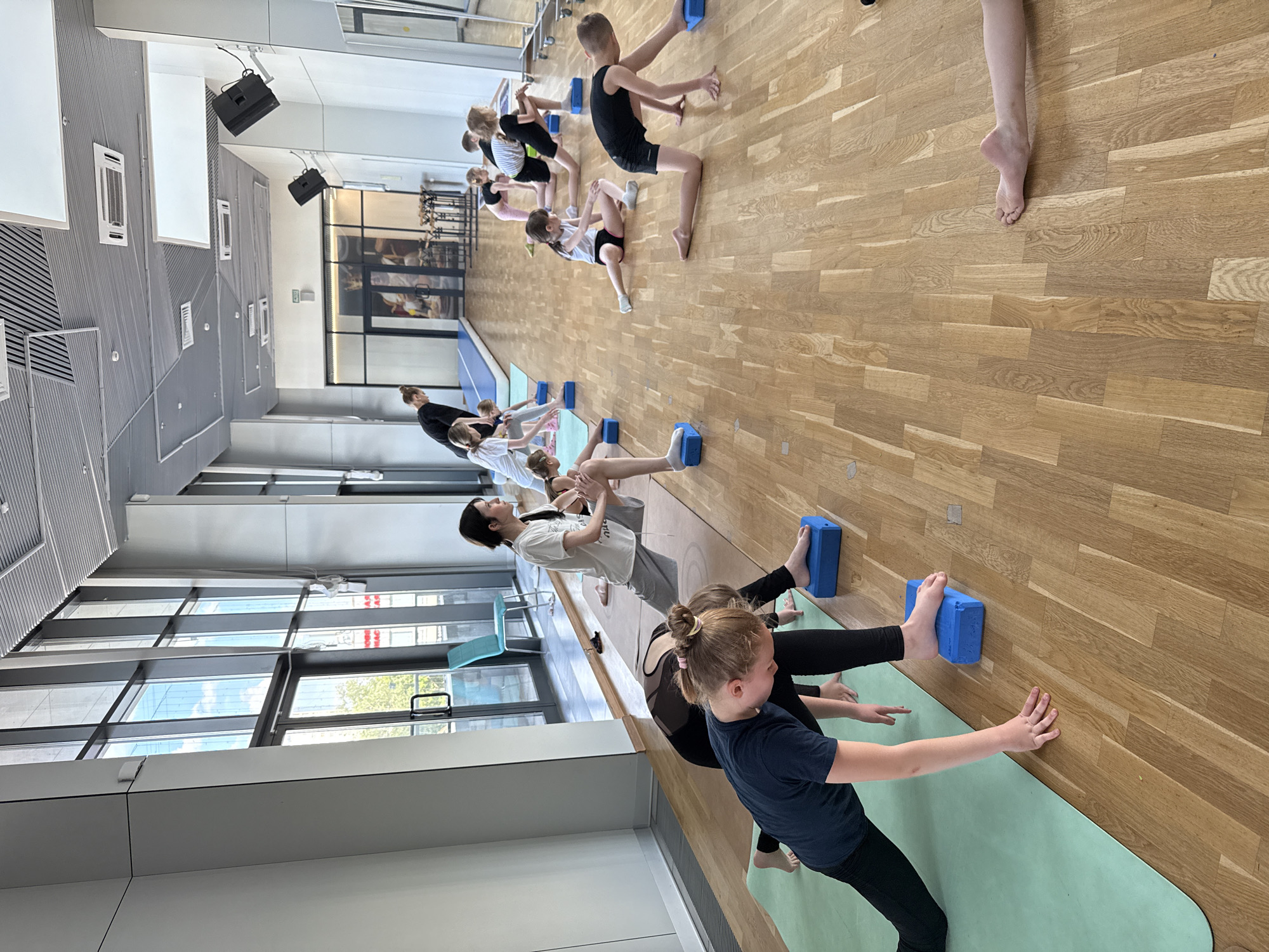 This image captures an indoor group exercise session, likely gymnastics or yoga, primarily involving children with a few adults. The setting is a bright, modern studio in Khimki, Russia, featuring a light wooden floor and light green exercise mats. Participants are engaged in various balancing and stretching poses, often utilizing blue foam blocks as props.
In the foreground, a young girl with a ponytail in a dark t-shirt and leggings is kneeling on a green mat, with her hands stretched forward towards the wooden floor, and one foot resting on a blue block. Behind her, another girl in black leggings is also using a blue block for support, while an adult, possibly an instructor, assists a child further back.
Towards the middle and background, several children, mostly pre-teens, are seen performing different exercises. Some are balancing on one leg with a foot on a blue block, while others are stretching or holding poses against the wooden floor. An adult, possibly an instructor with dark hair, is visible near the center, observing and guiding the participants.
The room is well-lit, suggesting daytime, with large windows along one wall revealing an exterior view of a city building and sky, reflecting some clouds. Mounted black speakers are visible on the ceiling, and air vents are integrated into the wall panels. The overall atmosphere appears focused and active. A small green "EXIT" sign is visible above a doorway in the background, and some red, reversed, and largely illegible text can be seen on a building through the windows, possibly "AC RAC".