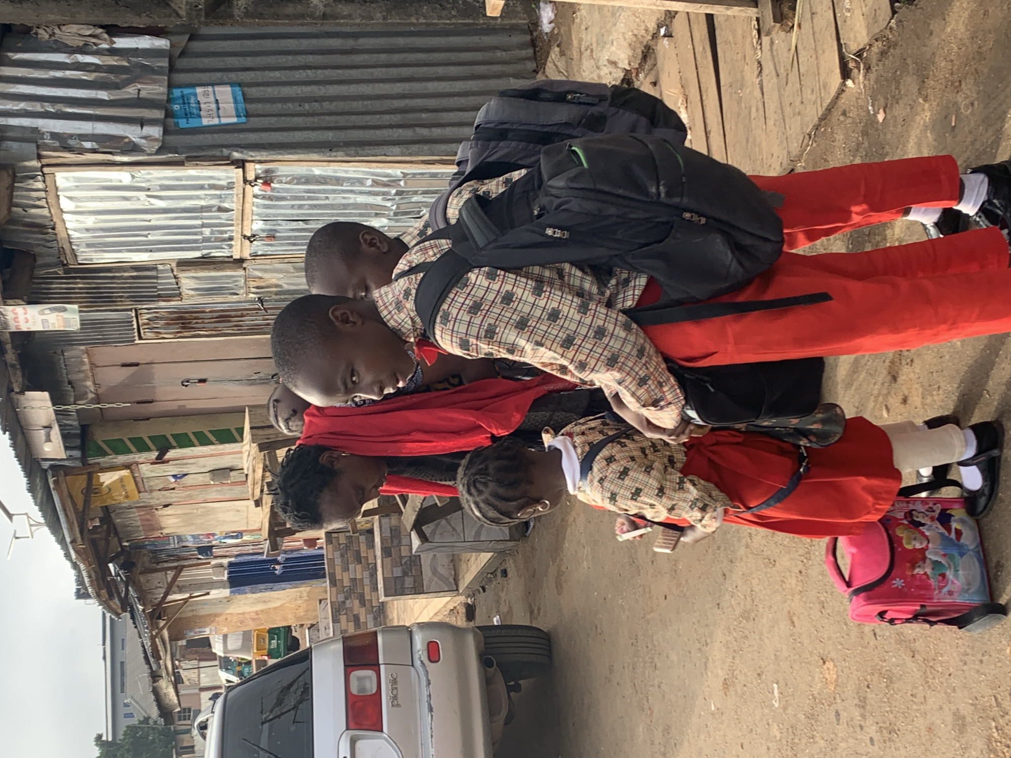 In the city of Jos, Nigeria, a group of three children, two boys and a young girl, stand with their mother. The children are dressed in school uniforms: the boys wear plaid shirts and red trousers, while the girl sports a plaid top and a red skirt. They are equipped with backpacks, and the girl also has a pink suitcase adorned with Disney princesses. The mother, wearing a red shawl, appears to be interacting with her children. The backdrop is a street scene characterized by informal shops constructed with corrugated metal and weathered wood. A silver car is parked to the left, and in the distance, a yellow tricycle is visible. The ground is unpaved, with scattered debris. The overall environment suggests a bustling, informal urban setting, likely a market area or residential street in Jos.