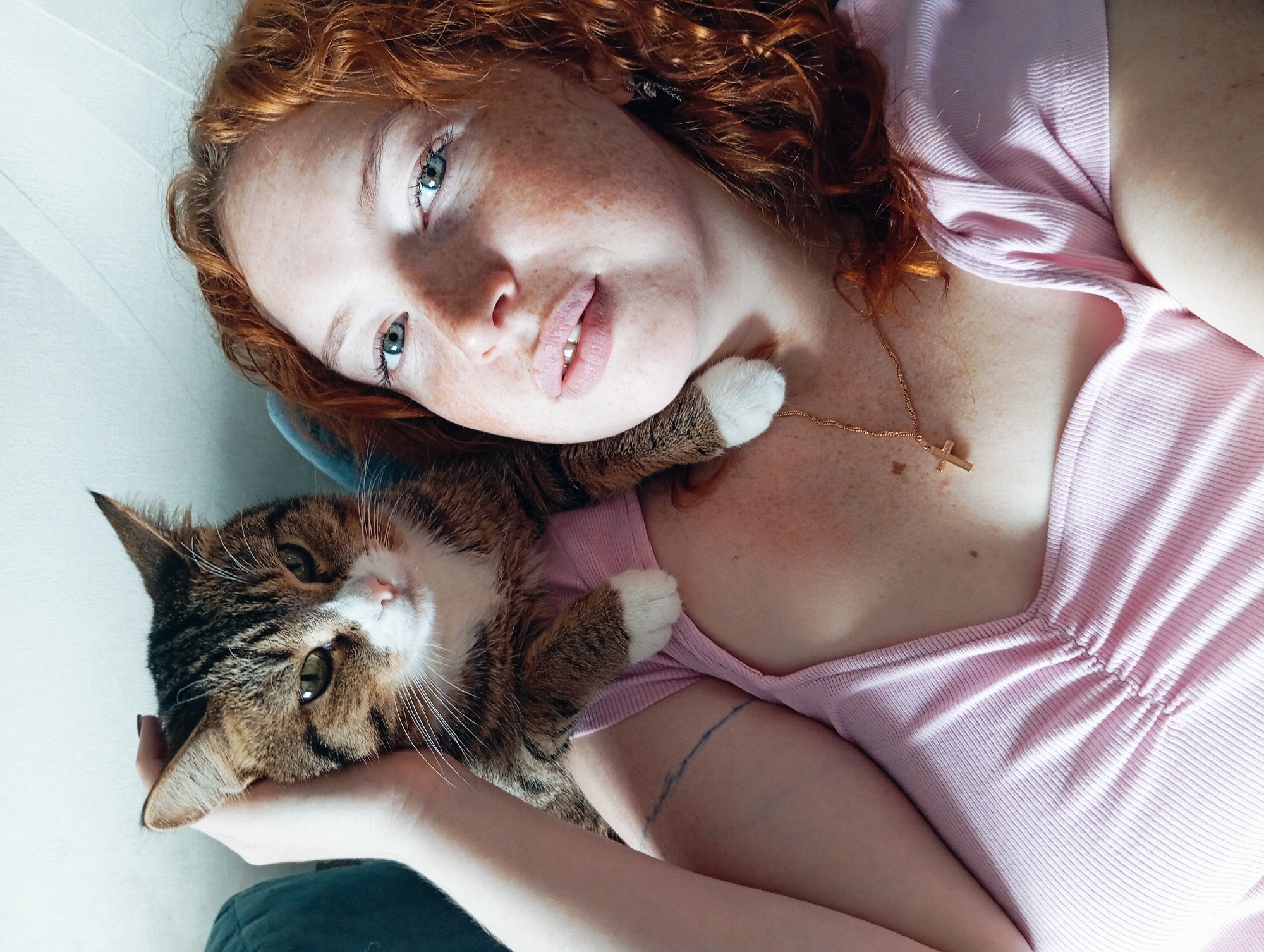 A fair-skinned woman with red curly hair and freckles is lying down with a tabby cat. The woman is wearing a light pink top and a gold necklace with a cross pendant. She is holding the cat with her left arm. The cat's head is resting on her arm, and its paws are visible on her top. The cat is a tabby with white markings on its face and paws. The background appears to be a wall and a cushion. The image was taken indoors, likely in a residential setting. The location of Yurgamysh, Russia is not evident from the image.