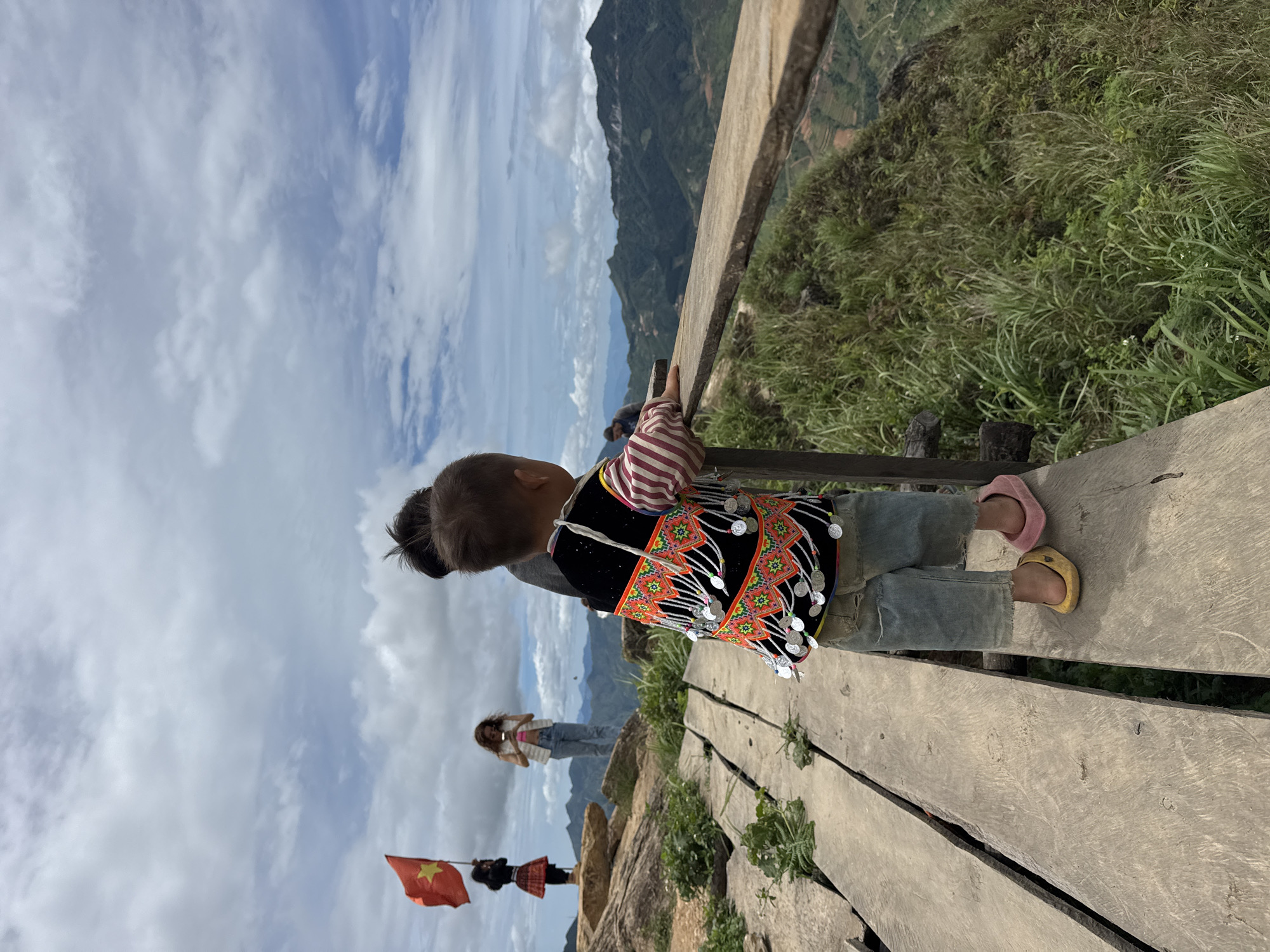 In Ta Xua, Vietnam, a young child, facing away from the camera, stands on a wooden walkway. The child wears a striped shirt and denim shorts, with a decorated vest adorned with silver coins and colorful embroidery. They are also wearing mismatched clogs, one yellow and one pink. The walkway extends along a steep, grassy hillside overlooking a mountainous landscape. In the background, two other individuals are visible on a rocky outcrop, one of whom holds a Vietnamese flag. The sky is partly cloudy.