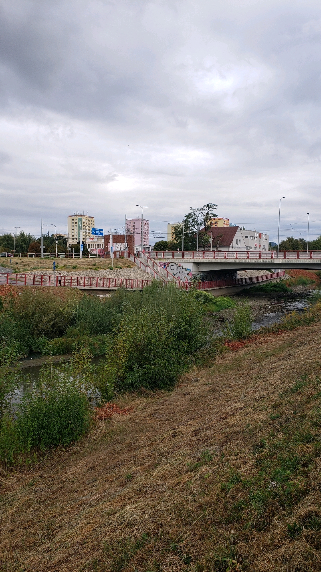 The image captures a view of a bridge over a river in the city of Prešov, Slovakia. The bridge, decorated with graffiti, connects two sides of the city, with a road running across it. Residential buildings and other structures are visible in the background. The riverbanks are lined with lush green vegetation, and dry grass covers the foreground. The sky is overcast, with dramatic clouds creating a moody atmosphere. The scene appears to be a typical urban landscape, with elements of nature and man-made structures coexisting.