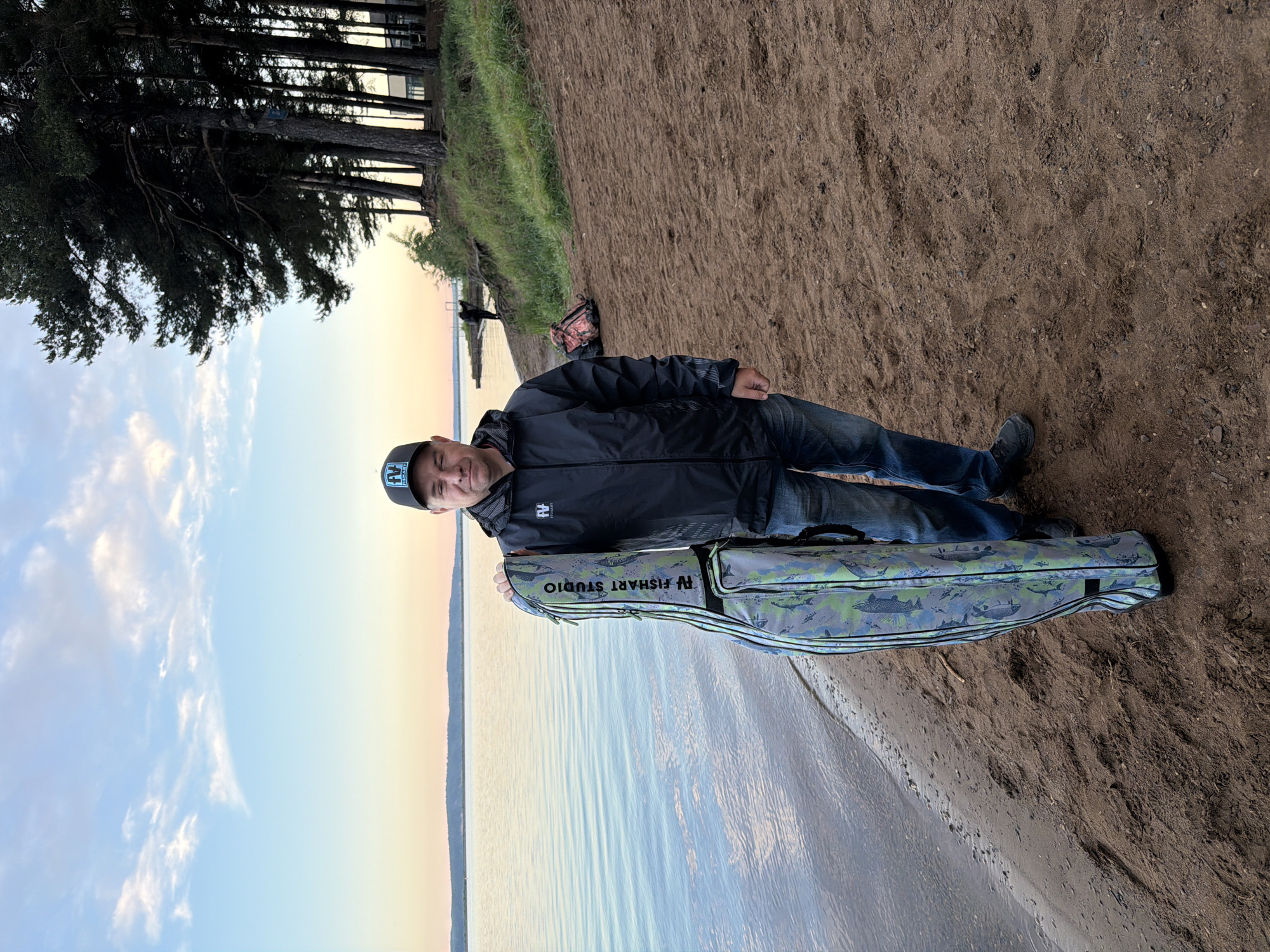 A man stands smiling slightly at the camera, posing on a sandy bank next to a calm body of water. He is wearing a black cap with a white "FV" logo, a black zip-up jacket with a matching "FV" logo on the left chest, blue jeans, and dark shoes. He is holding a tall, camouflage-patterned bag, likely a fishing rod case, which is adorned with various fish designs and prominently displays "FV FISHART STUDIO" in black text.
The setting is an outdoor landscape in Chaikovsky, Russia, characterized by a wide expanse of water, possibly a lake or river, extending into the distance under a vast sky. The sandy bank slopes gently down to the water's edge. To the man's left, a dense line of tall pine trees stands along the bank, with green grass and vegetation visible on the higher ground. Further down the bank, in the background, another person is partially visible near the water, with a dark bag or backpack lying on the sand.
The time of day appears to be either early morning or late evening, as indicated by the soft, warm light and the subtle hues of orange, pink, and light blue blending in the sky near the horizon, transitioning to a clearer blue with scattered white clouds overhead. The water's surface is calm, reflecting the sky's gentle colors. The scene suggests a tranquil outdoor activity, likely fishing or preparing for it.