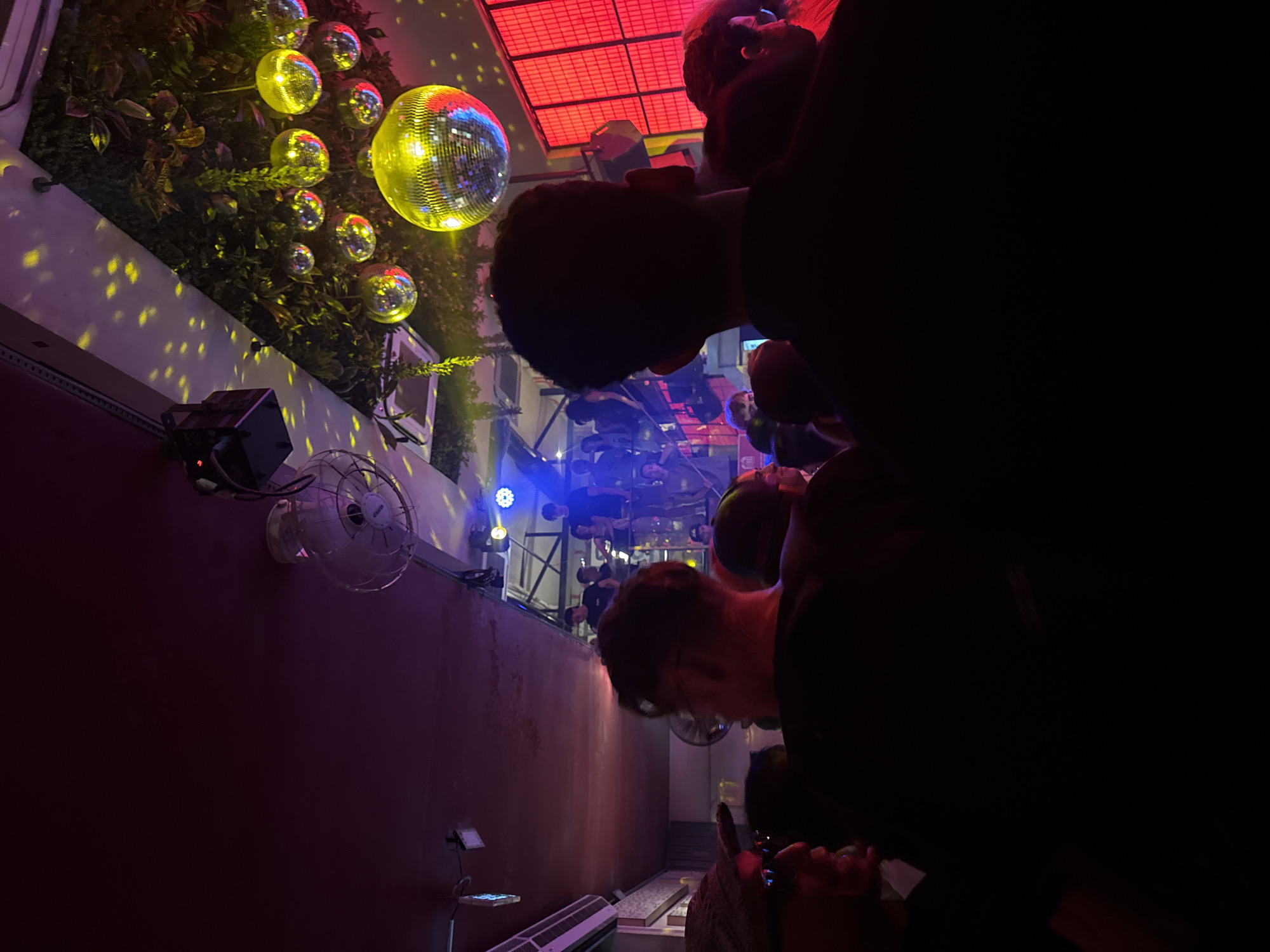 Captured in an unknown city, the image presents a lively nightlife scene. People, their features partially obscured by darkness, are visible towards the right side of the frame. Their silhouettes suggest they are standing close to one another, perhaps in conversation or observing the scene.
The environment is vibrant with multiple disco balls hanging from the top left, reflecting dynamic light patterns across the wall and nearby plants. A white wall-mounted fan adds a practical element to the space, possibly for ventilation.
In the background, a structural frame with blue and white spotlights illuminates the space, enhancing the ambiance. The red ceiling panels also contributes to the energetic atmosphere. The entire scene appears to be set in an outdoor venue or a rooftop bar, with a contemporary or trendy vibe.