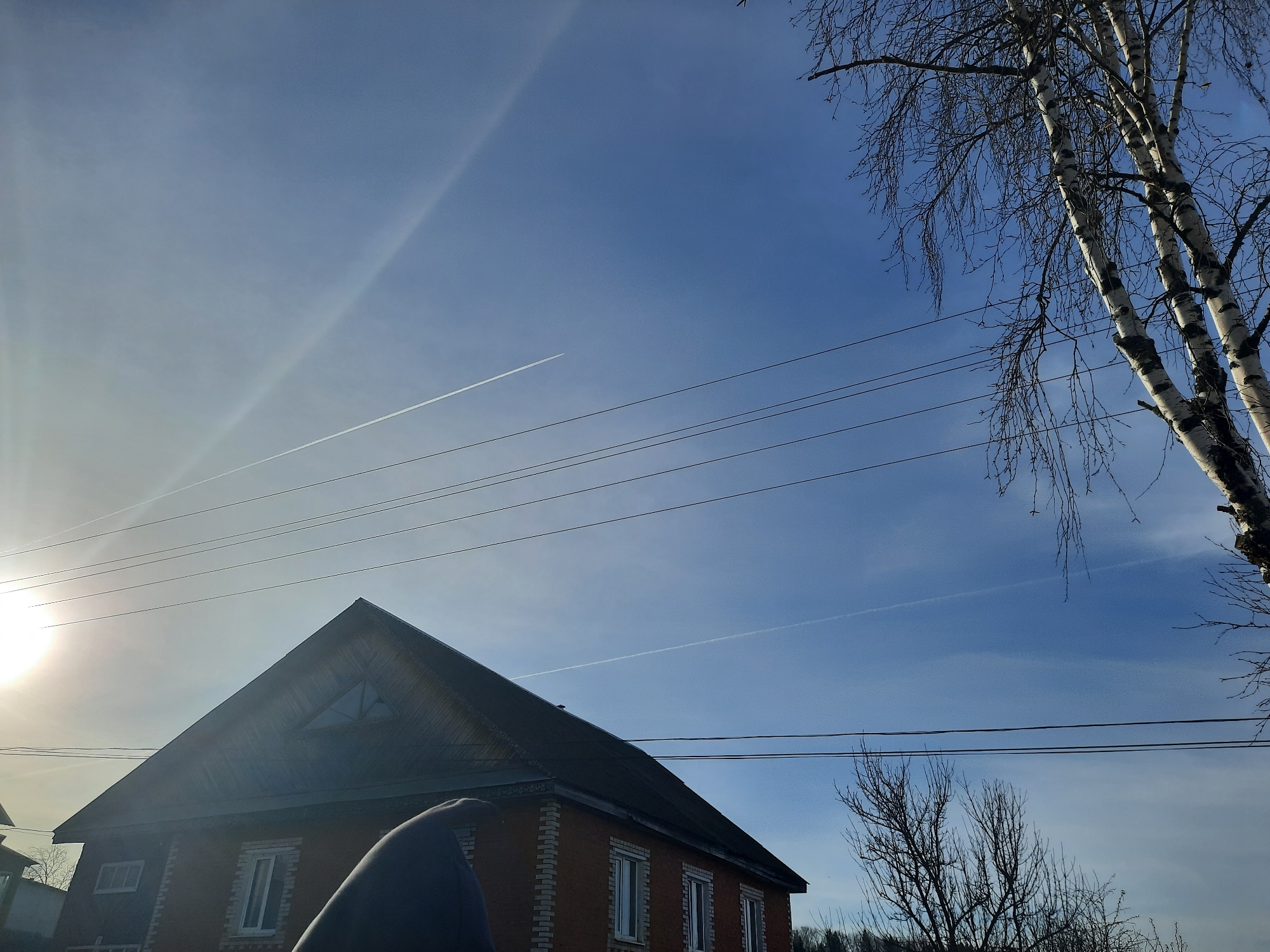 The image shows a two-story brick house with a dark roof in Krasnogorskoye, Russia. The house has multiple windows with white frames. Above the house, electrical wires stretch across the sky. The sky is blue with contrails. A birch tree with bare branches stands to the right of the house. The sun is visible in the lower-left corner, creating lens flare. The foreground features an out-of-focus blue object.