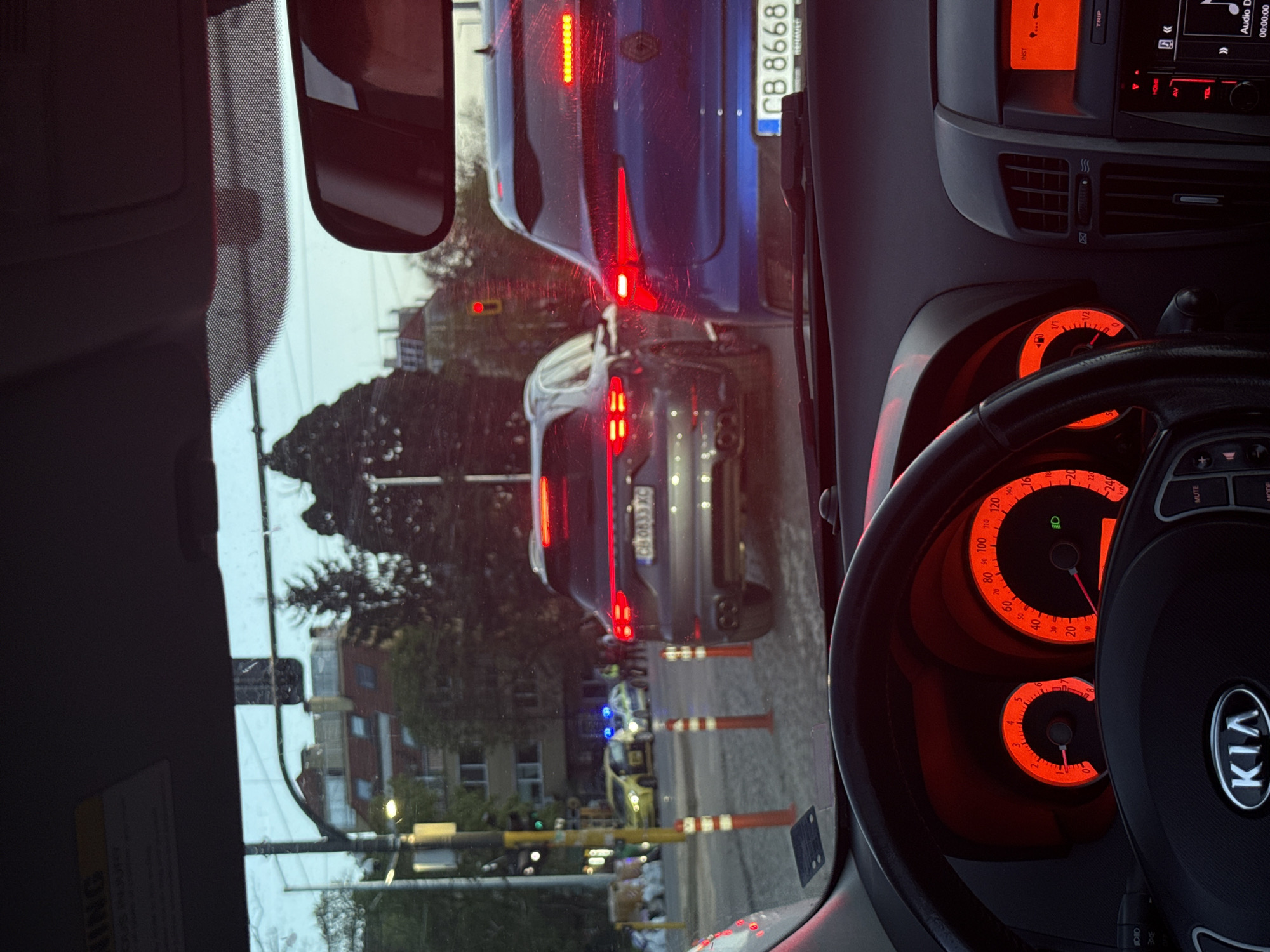 The image shows a view from inside a Kia car, looking out onto a street scene in Sofia, Bulgaria. The license plate of the vehicle in front of the Kia, a blue Renault, is "CB 8668 BJ." Behind the Renault is a grey car with the license plate "CB 0833 XC". There are striped safety bollards on the side of the road. In the distance, an ambulance can be seen with its emergency lights on. A traffic light is visible on the left. The car's dashboard is visible, illuminated in red. The steering wheel displays the Kia logo.