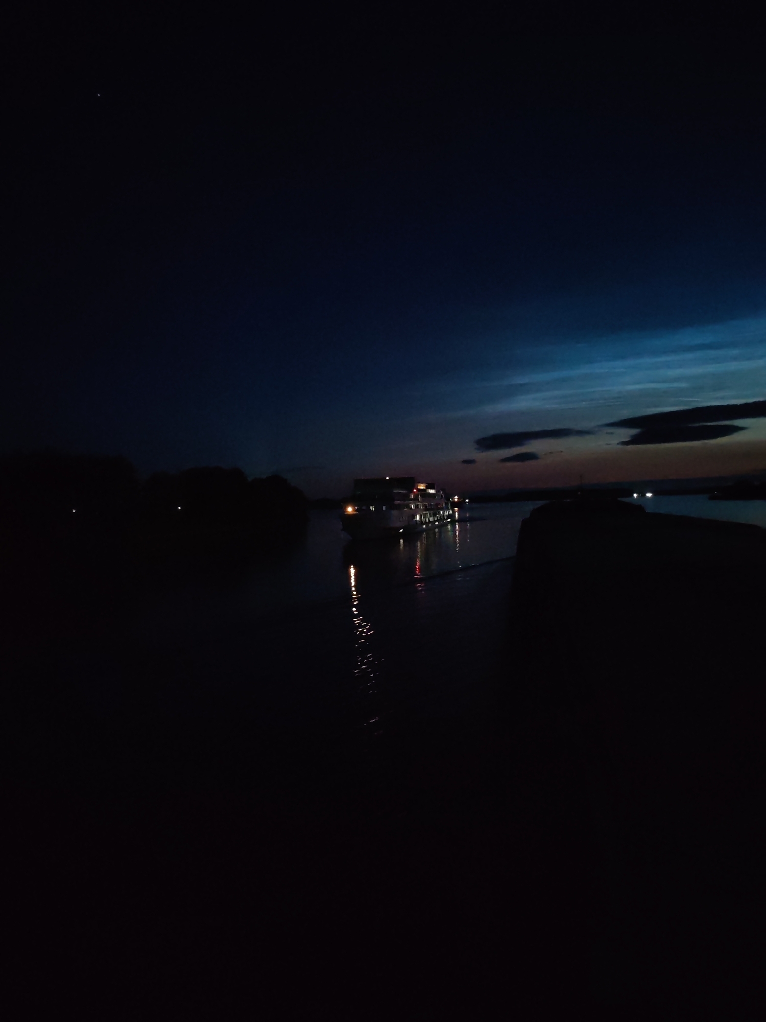 The image depicts a dark, twilight scene with a river and a boat. The sky is a deep blue, transitioning to lighter blues and grays near the horizon, with faint clouds visible. A single star is visible in the upper left quadrant of the image. On the left side of the river, silhouetted trees are visible, with a few faint lights scattered amongst them. A boat with illuminated windows is moving on the river, its lights reflecting on the water. The right side of the image is dominated by a large, dark, silhouetted object that appears to be a landmass or structure, possibly a pier or embankment. The overall impression is one of a quiet evening on the water. The location is identified as Bol'shie Mkhi, Russia.