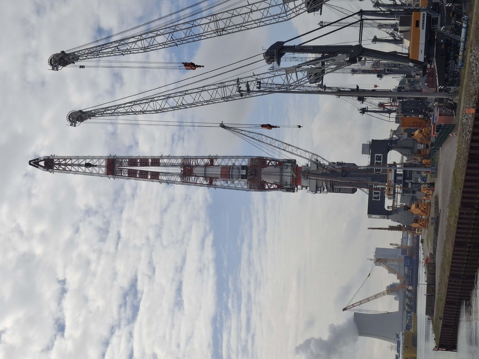 This media file presents a bustling industrial port or shipyard scene in Rostock, Germany, under a partly cloudy daytime sky. The primary subjects are multiple colossal industrial cranes, standing tall and prominent against the sky.
In the foreground, a striking red and white lattice-boom crane dominates the view, its massive structure extending diagonally upwards. Behind it, two more large cranes, primarily dark grey with yellow accents, are visible. The brand name "LIEBHERR" is clearly legible on the side of these two cranes, indicating their manufacturer. All cranes feature intricate metal lattice booms, extensive cable systems, and large hooks dangling, suggesting readiness for heavy lifting operations, though no active movement is depicted. Another partial text, "RR," can be seen on a structure closer to the water.
To the right of the image, a calm body of water, likely a harbor or canal, reflects the overcast sky. It borders a textured embankment, possibly made of concrete or sheet piles. On the land beyond the cranes and alongside the water, numerous industrial buildings, storage facilities, and other heavy equipment are scattered, reinforcing the industrial setting. The sky is a mix of bright white and grey clouds interspersed with areas of clear blue. No people are visible in the frame, allowing the immense scale of the machinery and the infrastructure to take center stage. The overall impression is one of substantial industrial activity and capacity.