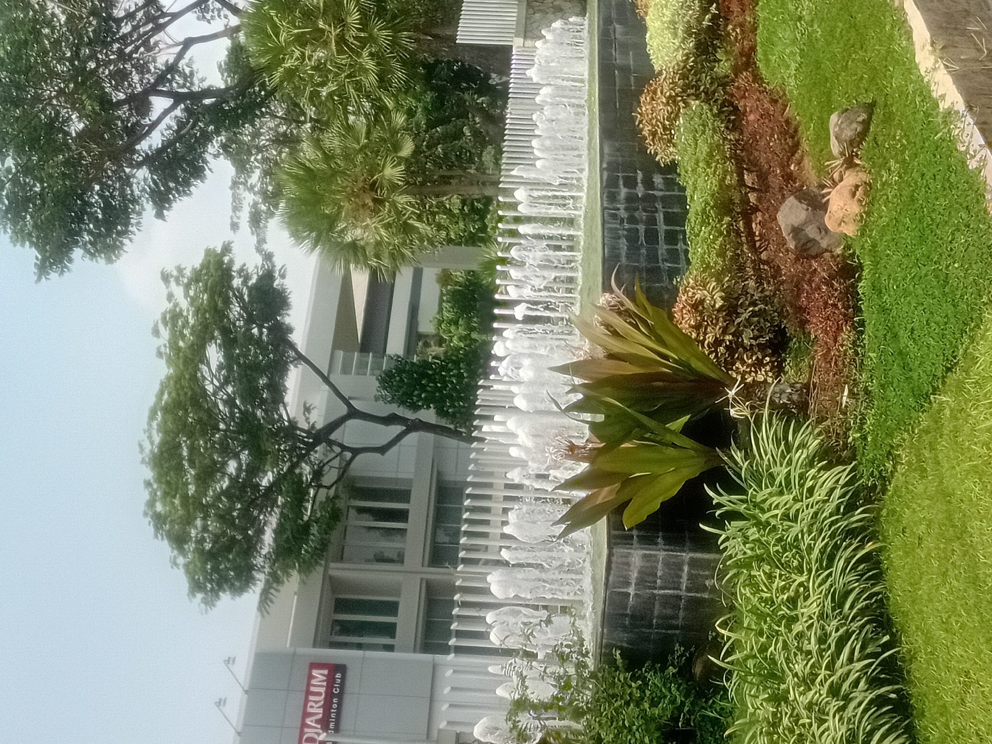 The image shows an outdoor scene featuring a fountain in front of a modern building with the sign "DIARUM Badminton Club". The fountain has multiple streams of water spraying upwards, creating a lively effect. Lush green grass covers the foreground, with manicured bushes and plants surrounding the fountain. In the background, palm trees and other foliage add to the tropical ambiance. The building appears to be well-maintained, suggesting it is an active sports facility. The sky is bright, indicating a sunny day. The scene suggests a public space, possibly a park or a recreational area within Klentengan, Indonesia, designed for leisure and sports activities. There are no people visible in the image.