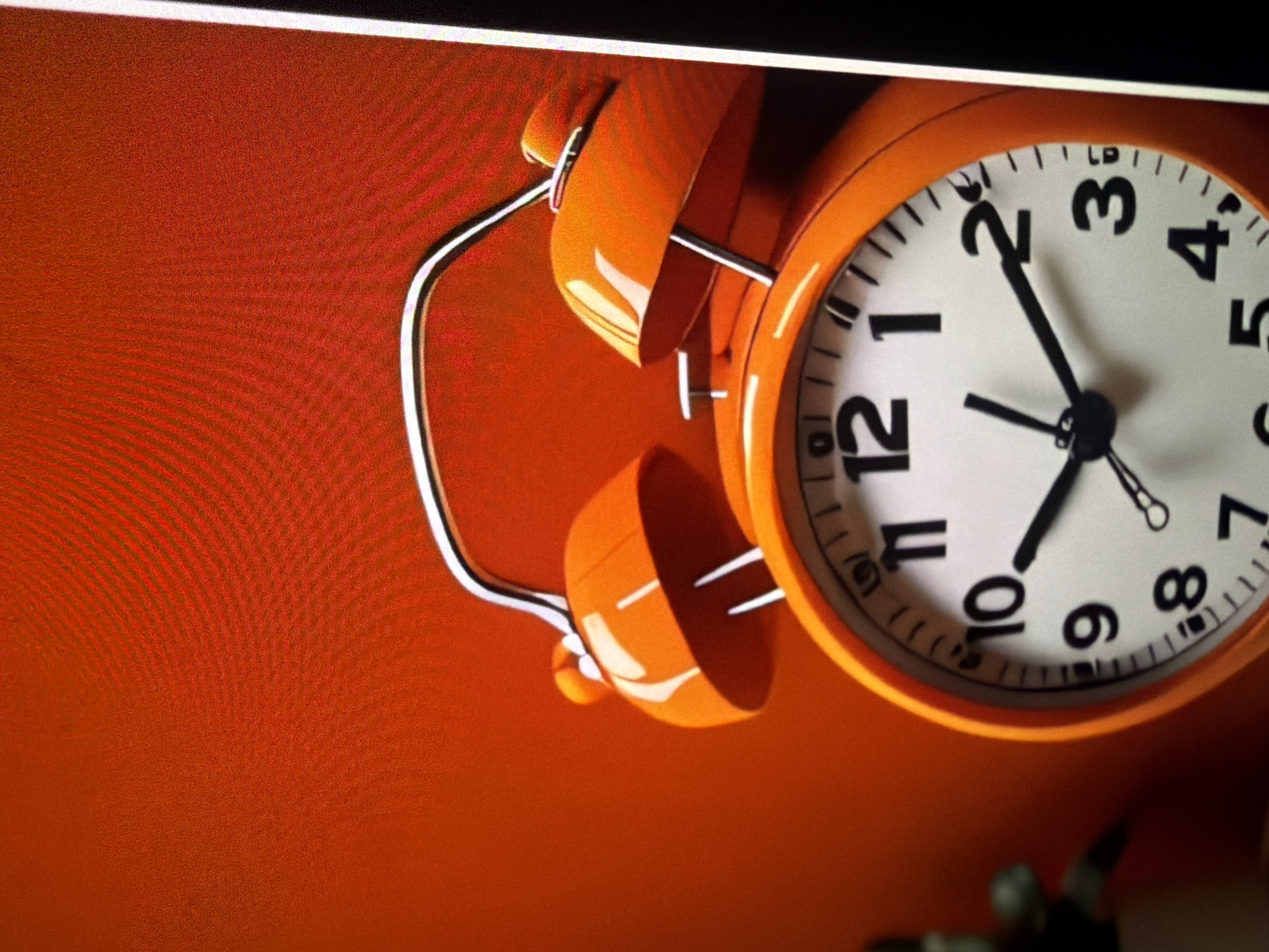 The image shows a close-up of an orange analog alarm clock against a textured red background. The clock features black Arabic numerals for hours 1 through 12, and black hands indicating the time. The hour hand points between the 9 and 10, and the minute hand points towards the 2. The clock has two bells on top, characteristic of a twin-bell alarm clock, and a silver metal handle connecting them. The lighting in the image creates subtle reflections on the glossy surface of the clock. To the left of the clock, a blurred green object, possibly a plant, is partially visible. The scene is indoors, with no indication of a specific location in a city.
