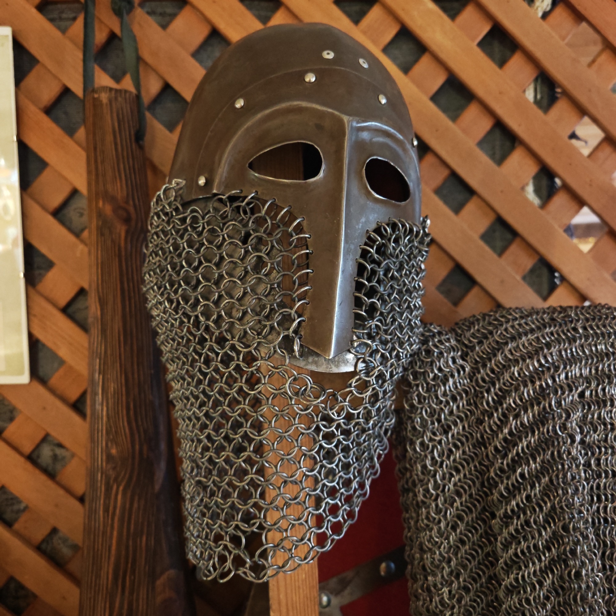 A medieval helmet with attached chainmail covering the face is positioned against a wooden lattice background. To the left of the helmet is a long, narrow piece of dark brown wood. To the right, more chainmail is visible, partially obscuring a red surface with metal detailing. The image was taken in the city of Rostov, Russia.