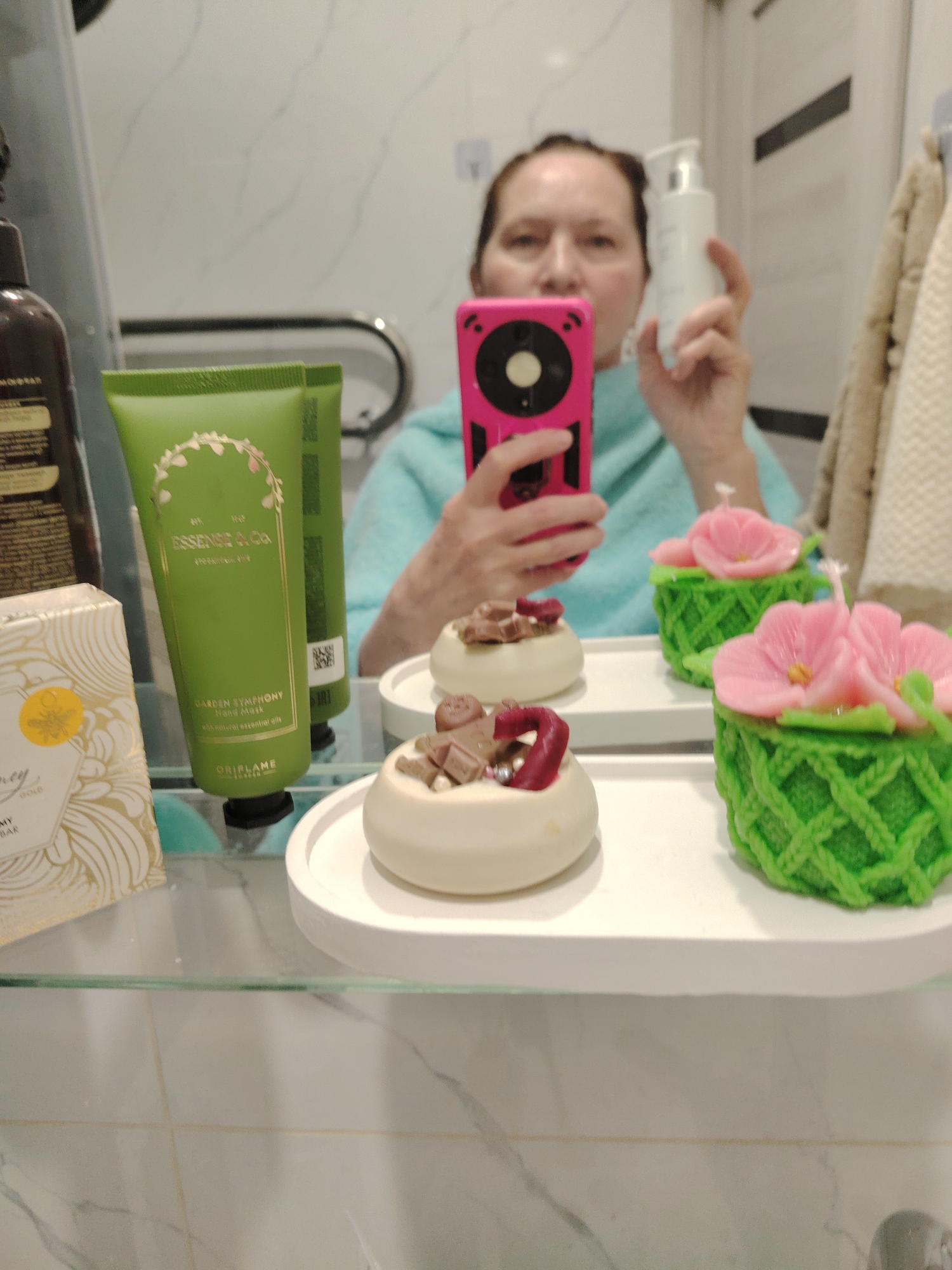 The scene is captured in a bathroom, possibly within a residence in Sarapul, Russia. A person is taking a selfie, holding a pink smartphone, and partially obscured behind the phone is a person holding a white bottle. In the foreground, arranged on what appears to be a glass shelf, are several items. These include a box of soap, a green tube of hand cream from the "Essense & Co" line by Oriflame, and two decorative candles shaped like small cakes, one topped with pink flowers and the other with chocolate-like pieces. The background shows a bathroom with a white wall, a towel, and a metallic handrail.