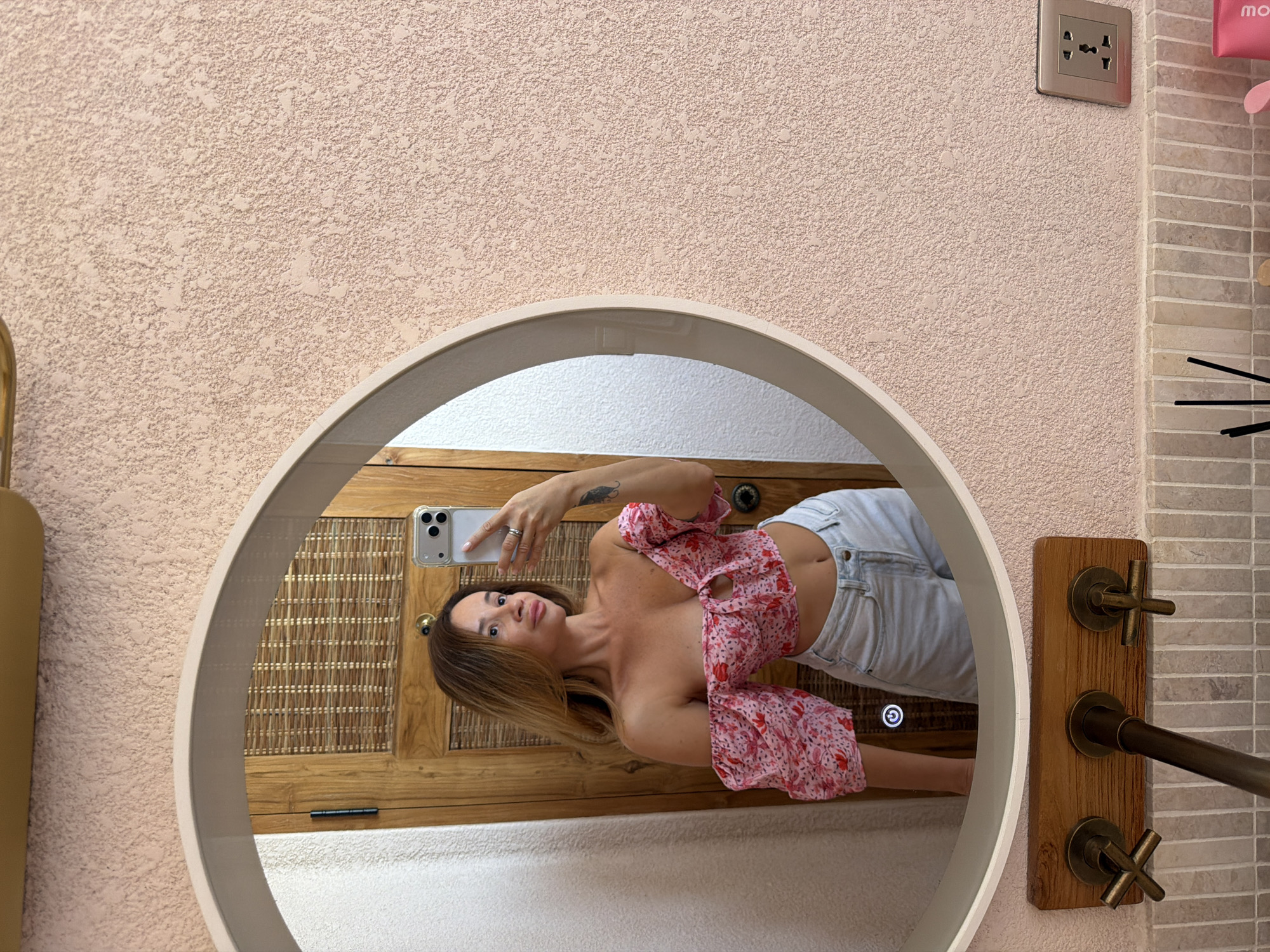 A woman is taking a mirror selfie in what appears to be a bathroom in Mengwi, Indonesia. She is wearing a pink floral off-the-shoulder crop top with puff sleeves and light wash denim shorts. The mirror is round and framed by a beige border. The wall behind her is textured and painted a light pink. The background reflected in the mirror shows a wooden door with woven details. Below the mirror, there is a wooden fixture with two cross-shaped faucets and a central pipe. The wall beneath this fixture is tiled with small, light-colored rectangular tiles. To the right of the faucets, an electrical outlet with multiple sockets is visible on the pink textured wall.