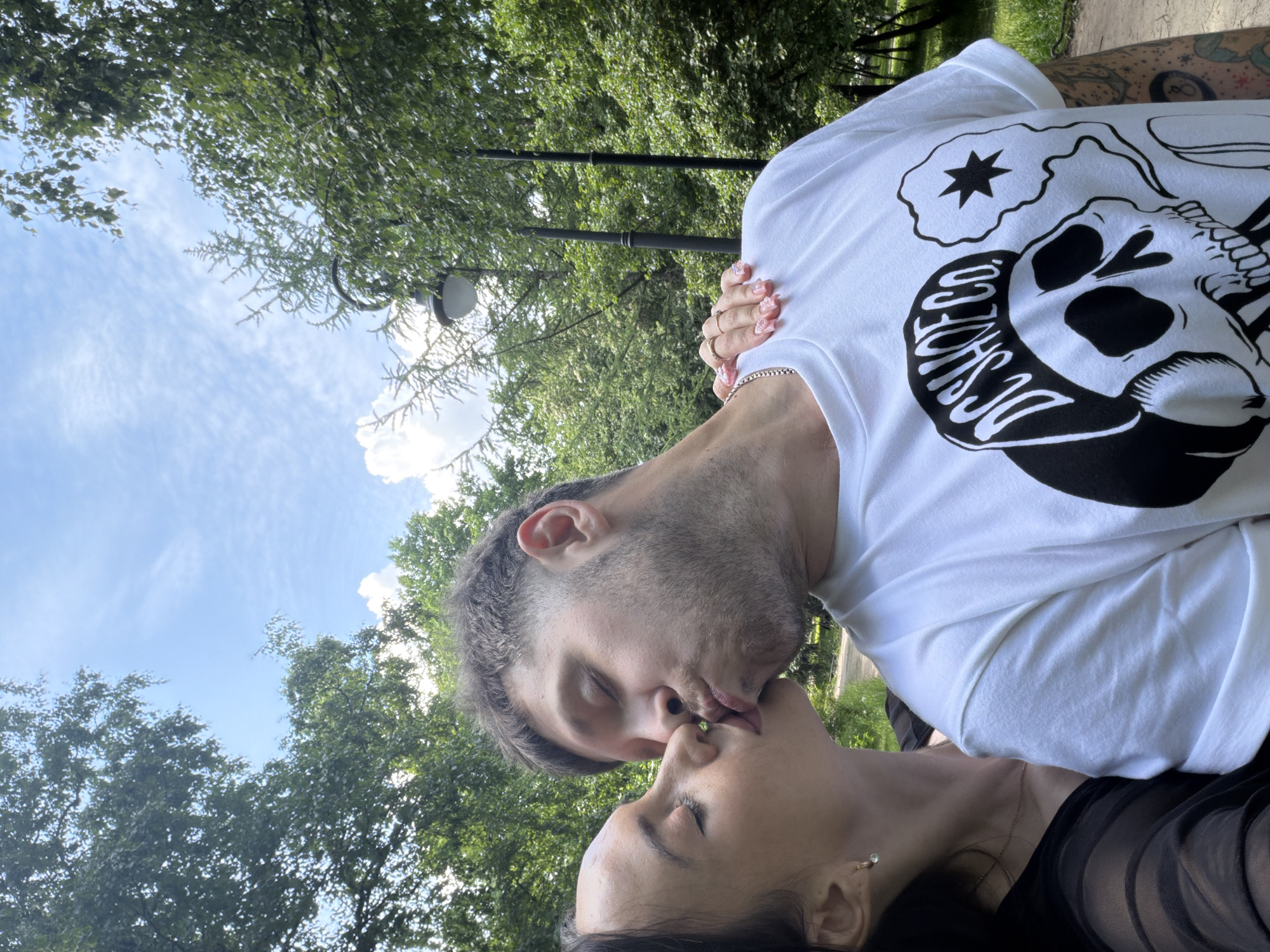 The image displays a close-up, chest-up view of two individuals, one female and one male, engaging in a kiss in an outdoor park environment. The female, positioned on the left, has dark hair and wears a sheer black top. She is seen with a gold hoop earring featuring a clear stone and a thin gold necklace. Her right hand, adorned with multiple rings on the middle and ring fingers and long, light pink artificial nails, rests on the male's left shoulder. The male, positioned on the right, has short dark hair with faded sides and a beard. He wears a white short-sleeved t-shirt with a prominent black graphic of a skull wearing a cap labeled "DC SHOE CO." and a thought bubble containing a black star. A silver chain is visible around his neck, and tattoos, including an "8-ball" design, are visible on his left arm. Both individuals have their eyes closed, with their lips touching, and the female's tongue is slightly visible.
The background features dense green foliage from trees and bushes, suggesting a park or wooded area. The sky above is blue with scattered white, wispy clouds, indicating a bright, sunny daytime setting. A black lamppost with a white fixture is visible behind the male's head on the right side of the frame, with another dark pole further in the background. A dirt or gravel path is faintly discernible in the distant background on the right. No specific landmarks are present to definitively identify the exact location within Saint Petersburg, Russia; however, the general park environment is consistent with urban green spaces found in the city.