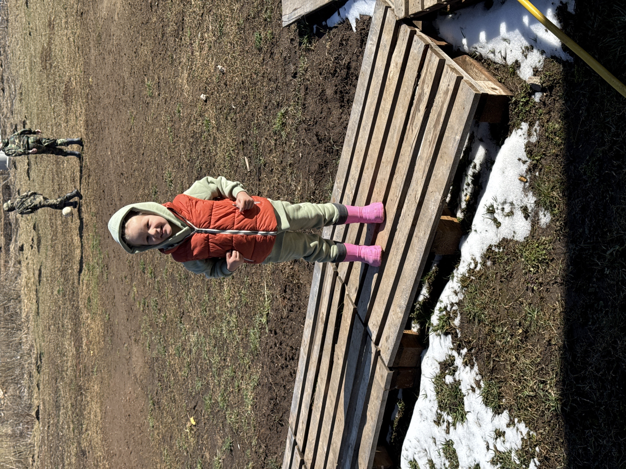 The scene depicts a child in pink boots, a green sweatsuit, and a red vest lying on a wooden pallet in an outdoor environment. Patches of snow are visible on the ground adjacent to the pallet, suggesting early spring conditions. In the background, there's a field or grassy area and two figures in camouflage attire. The photograph appears to have been taken in Gornyi, Russia.