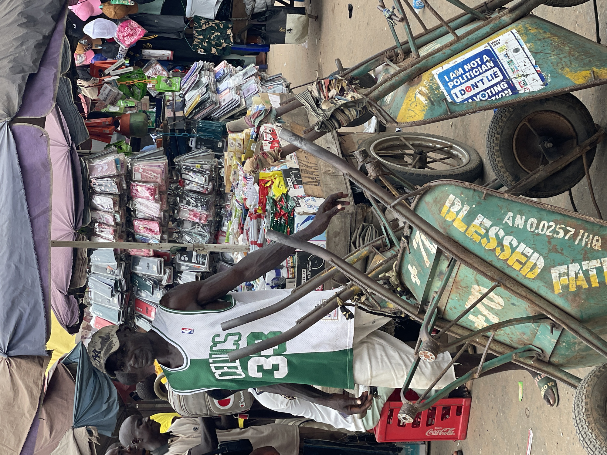 The image depicts a street scene in Onitsha, Nigeria. A man wearing a Boston Celtics jersey and a camouflage cap stands amidst a bustling market. A variety of merchandise, including phone cases, clothing, and other items, are displayed on makeshift tables. A pair of heavily-used, green hand trucks with slogans painted on them, including "BLESSED FATT" and "I AM NOT A POLITICIAN I DON'T LIE," are positioned nearby, one holding a red Coca-Cola crate. Other individuals can be seen partially in the foreground. The setting appears to be a crowded commercial area with merchandise overflowing onto the walkways.