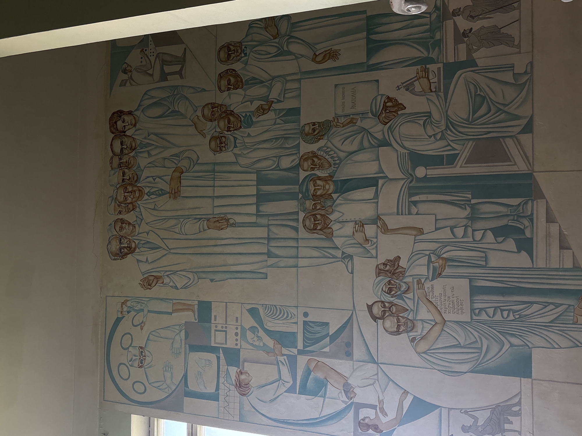 The image shows a mural depicting medical figures and scientific concepts in Perm, Russia. The mural is on a light-colored wall. The style is stylized, with the figures having a somewhat Byzantine-like appearance.
The mural contains multiple sections featuring different scenes. The top section features a series of men in white coats, possibly doctors or scientists. The middle section contains figures in robes. One holds a book with the words "VESALIUS ANDREAS ANATOMIA" and other Greek text that includes words such as "πλευμονία" (pneumonia) and "φθίσις" (phthisis - tuberculosis). The bottom section includes images of medical procedures, anatomical representations, and other diagrams. There is a figure lying down with images of a skeleton above.