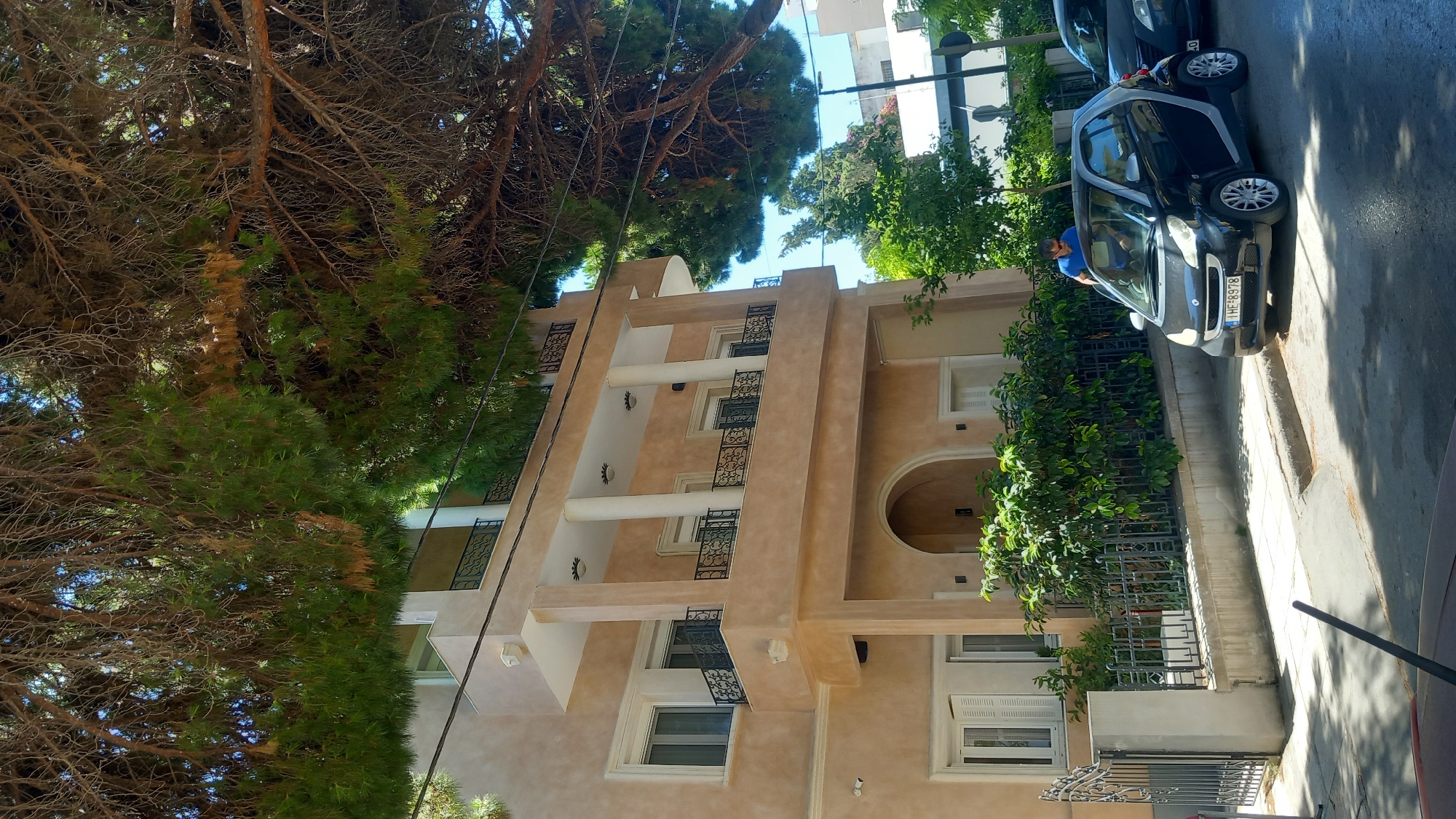 An outdoor shot captures a scene on a street in Kos, Greece, with a multi-story building and parked cars. In the foreground, a black Smart car is parked on the right side of the road. A man wearing a blue t-shirt is visible, leaning into the driver's side of the Smart car. Behind the Smart car, a portion of another dark-colored vehicle is visible. The building, painted in a light beige color, occupies the background and features balconies with decorative railings and pillars supporting an overhang. Lush green foliage and trees, including pine trees with their branches extending over the building, are present throughout the scene. The sky is a clear blue. The overall environment suggests a sunny day in a residential or commercial area.