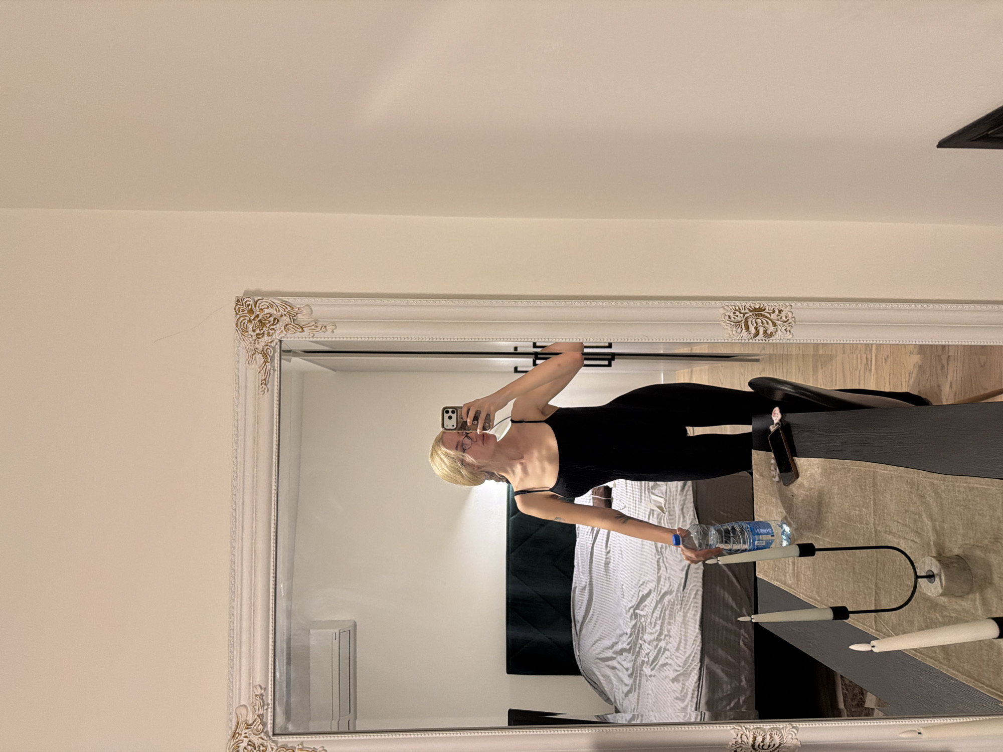 A mirror selfie shows a woman in a black outfit holding a water bottle. She is standing in a bedroom with a bed and a bedside table that holds a modern candle holder. The environment suggests a residential setting. It is not possible to determine if this is in Belgrade, Serbia from the image alone.