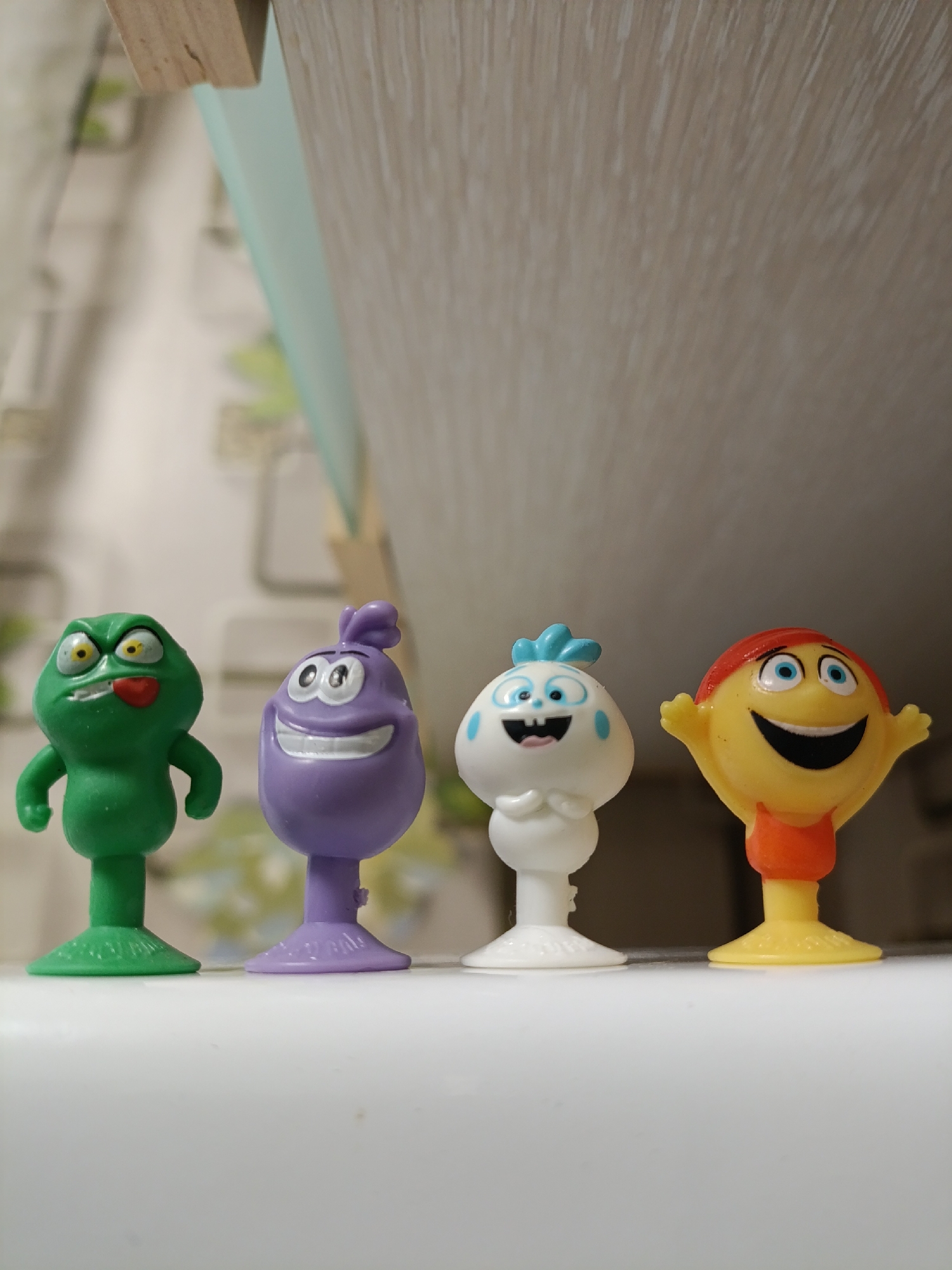 Four small, colorful toy figures stand in a row on a white surface. From left to right, the figures are: a green frog-like creature with bulging yellow eyes and its tongue sticking out; a purple blob-like creature with wide eyes and a large grin; a white, round creature with blue accents and a small mouth; and a yellow, round creature with blue eyes, a wide black mouth, and an orange collar. The figures appear to be made of plastic and have individual bases. The background is blurred, showing a light-colored textured surface and a hint of greenery. The lighting suggests an indoor environment.