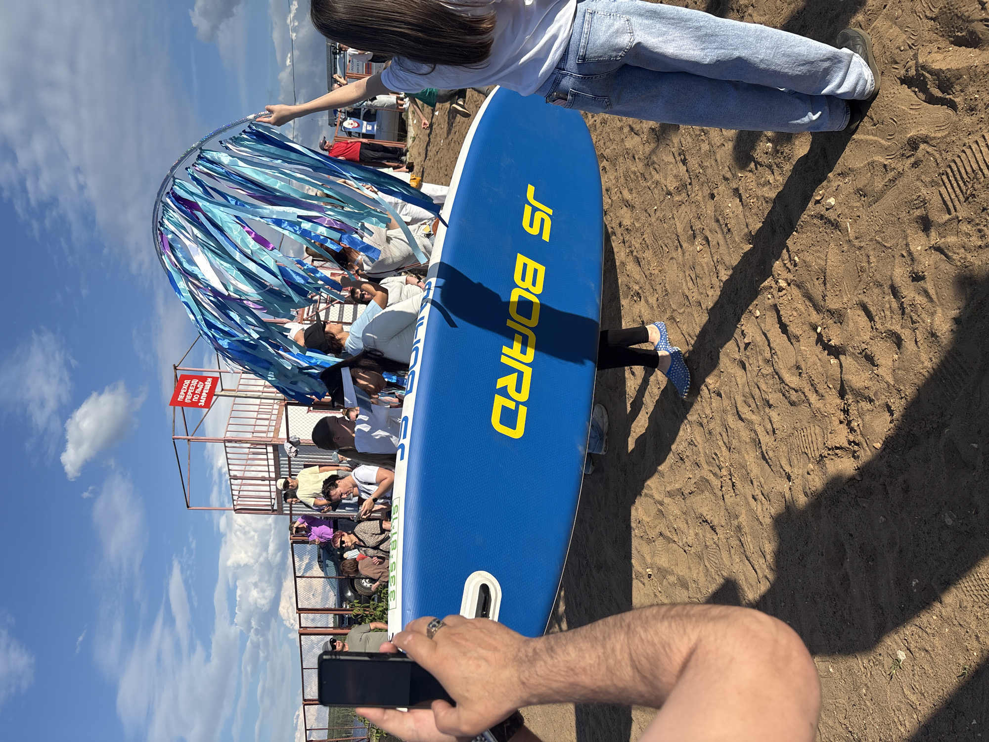 A sunny day in Chaikovsky, Russia, features a group of people gathered on a sandy area. The central focus is a person holding a blue and yellow paddleboard, with the brand "JS BOARD" prominently displayed in yellow lettering. Behind the paddleboard, a large, hoop-like structure adorned with blue and purple streamers is being held up by an individual. In the background, a red and white metal structure, possibly a viewing platform or part of a sports facility, stands against a clear blue sky with scattered clouds. Several individuals are visible, some holding phones, suggesting they are documenting the event. A red sign with white Cyrillic lettering is affixed to the metal structure. The ground is covered in sand, with tire tracks visible, indicating vehicular activity. The overall scene suggests a recreational event or gathering.