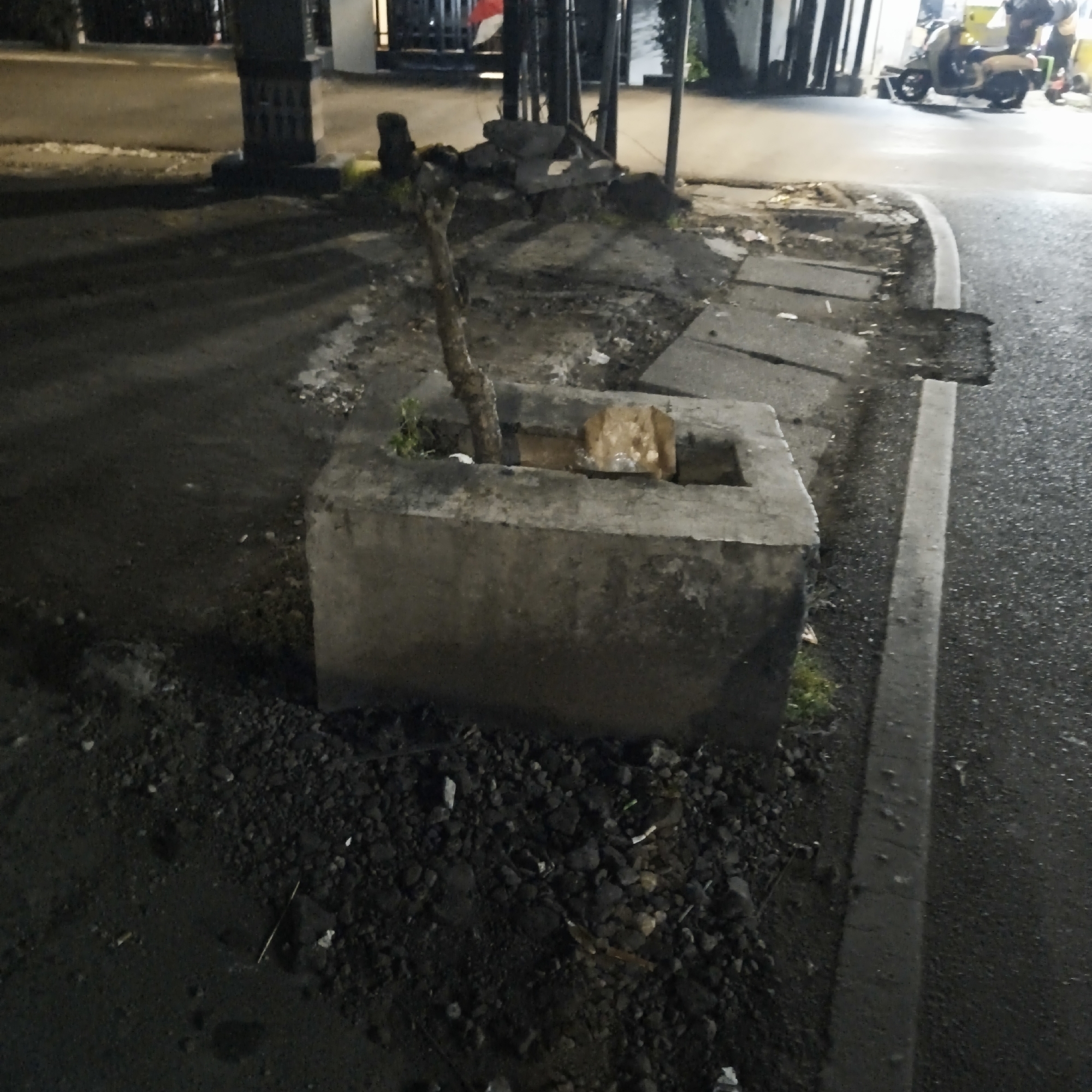 The image depicts an outdoor scene at night, likely in an urban environment. In the foreground, there is a concrete planter with a small, dead tree growing out of it. Some debris, including bricks and rocks, is scattered around the planter and on the surrounding ground, which appears to be a mixture of dirt, gravel, and asphalt. To the right, a paved road with a concrete curb is visible. In the background, there are some buildings and a motorcycle with a person on it. The lighting suggests artificial light sources are present, creating shadows and illuminating parts of the scene. The overall impression is one of a somewhat neglected urban space at night. The location provided in South Jakarta, Indonesia, contextually places this scene within a tropical urban setting.