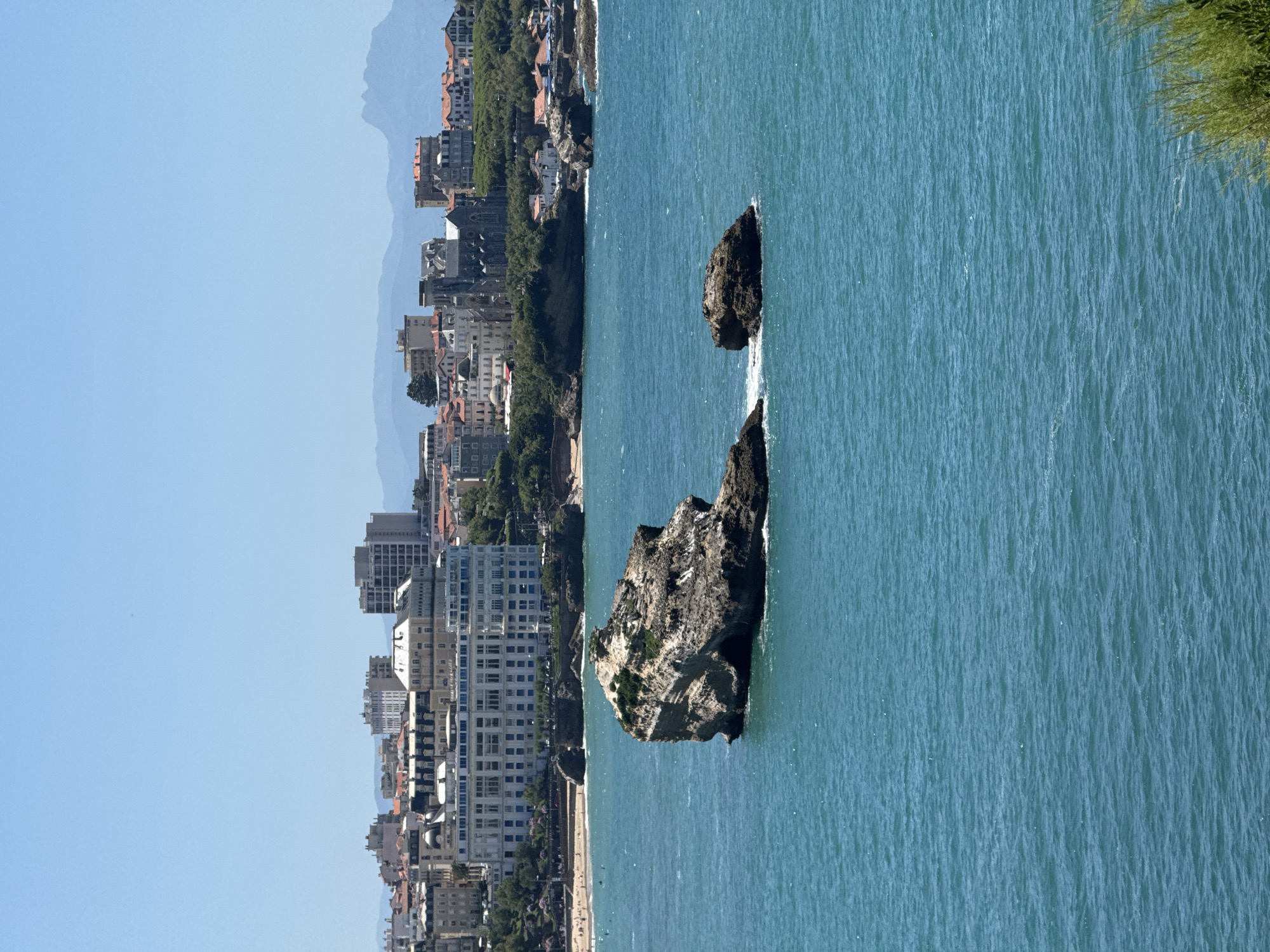 The image displays a coastal view of Biarritz, France, focusing on the iconic Rocher de la Vierge (Rock of the Virgin). In the foreground, the deep blue-green water of the Atlantic Ocean is visible, with choppy waves creating ripples on the surface. Two prominent rock formations rise from the sea. The larger rock, positioned slightly to the left of center, is rugged and layered, with patches of green vegetation growing on its upper surface. A smaller rock lies to its right, also partially submerged.
In the midground, a sandy beach is visible, dotted with a few people. Behind the beach, a picturesque cityscape unfolds. A large, white, multi-story building with a classical architectural style dominates the skyline, its numerous windows reflecting the bright sunlight. This building appears to be the Hôtel du Palais. To its left and right, other buildings of varying heights and architectural styles, including red-tiled roofs, form a dense urban landscape. A church with a tall spire is also discernible within the city.
The background features a clear, light blue sky. In the far distance, hazy mountains rise, indicating the presence of inland terrain. The overall scene is bathed in bright daylight, suggesting a warm, sunny day. The image captures the confluence of natural coastal beauty and the architectural charm of Biarritz.