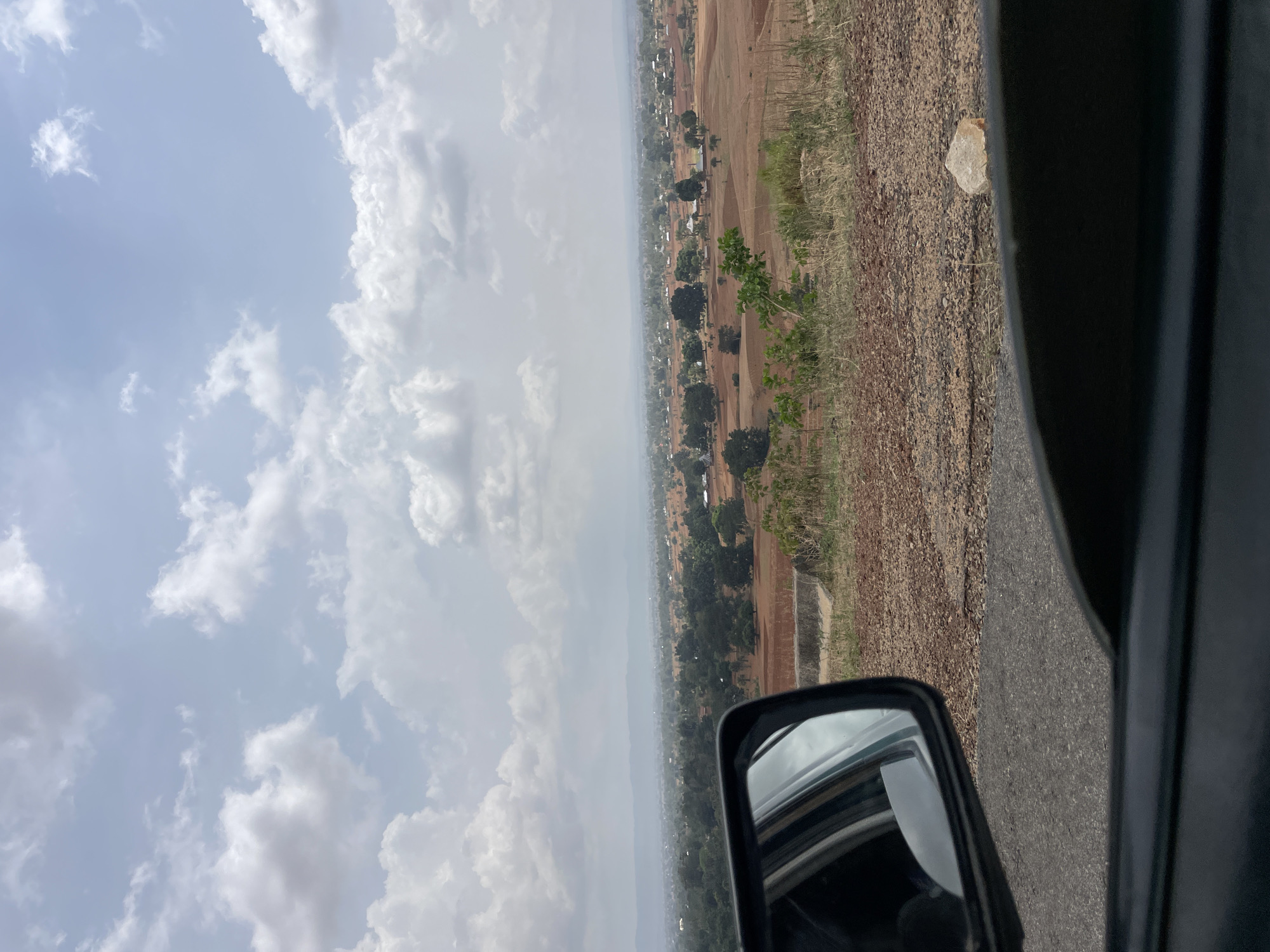 The image shows a view from inside a moving vehicle. A side mirror is visible in the foreground. Beyond the car's window, the scene unfolds onto a landscape featuring a cloudy sky above a rural landscape. In the distance, a settlement of Vom, Nigeria, comprising scattered buildings and trees, stretches across a flat, arable land, appearing reddish-brown. A road surface is visible immediately next to the car. There is also a close up shot of a rock next to the road.