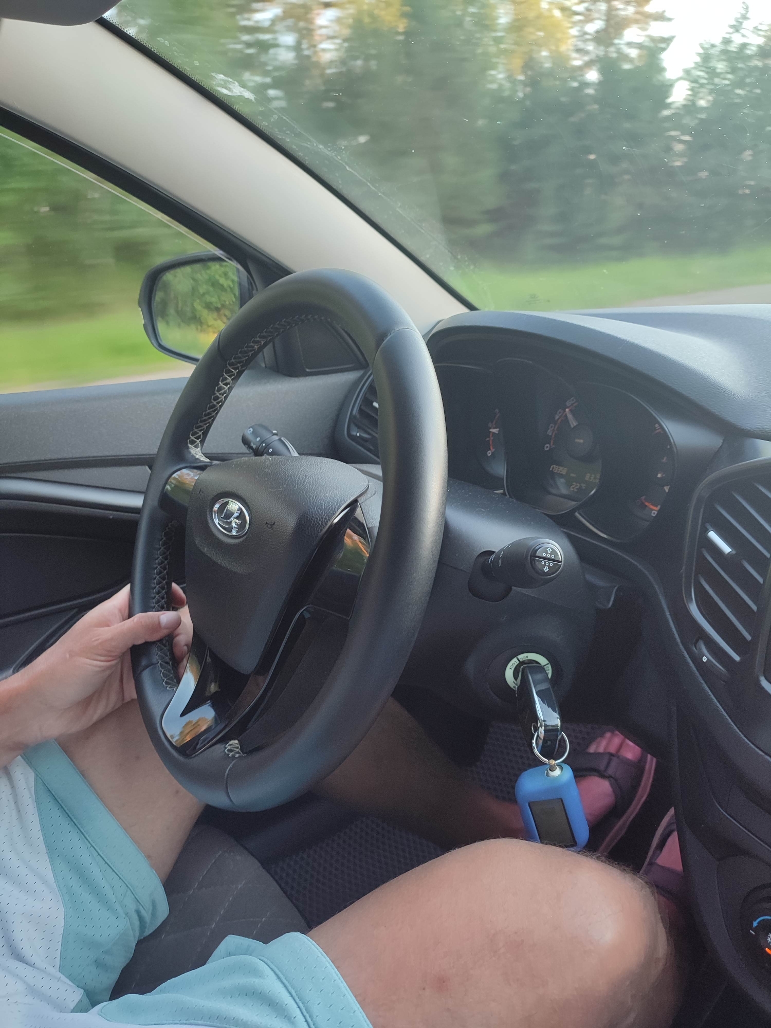 A first-person perspective captures a driver's hands on the steering wheel of a car. The driver, wearing a light-colored t-shirt with aqua accents and aqua shorts, is gripping the steering wheel. The car's dashboard is visible, including the steering wheel with a central logo, a turn signal lever, and the instrument cluster displaying speed and temperature. A blue car key fob hangs from the ignition. Outside the car, green trees and foliage are visible through the windshield, suggesting a drive through a natural setting. The scene appears to be taking place during the day, with soft lighting filtering through the trees.