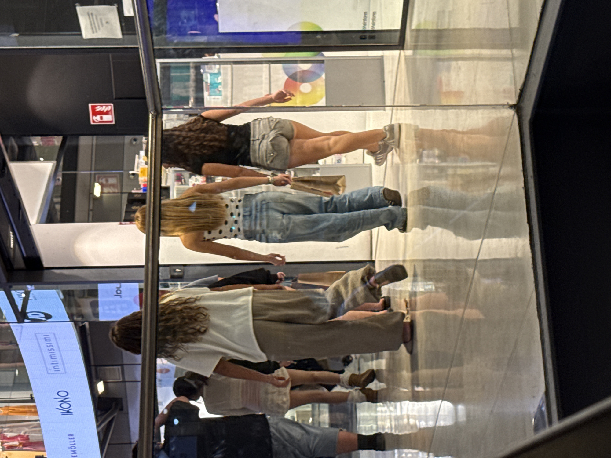 A group of young women walk through a brightly lit indoor shopping mall with polished floors that reflect the surroundings. The women are walking away from the camera, with their backs to the viewer. One woman in the foreground wears a white t-shirt and loose, tan pants. To her left, a woman in a light-colored top and a white skirt walks. To her right, a woman with long, dark curly hair wears a black t-shirt and denim shorts. Further to the right, another woman with long blonde hair wears a white top with a polka-dot pattern and blue jeans. A fire extinguisher sign is visible on a black wall in the background. The mall appears to be modern, with large glass storefronts and a digital display board overhead that reads "EMÖLLER IKONO intimissimi". The scene is captured in a medium shot, focusing on the lower bodies and backs of the individuals. The overall impression is one of casual shopping activity. This scene is located in Barcelona, Spain.