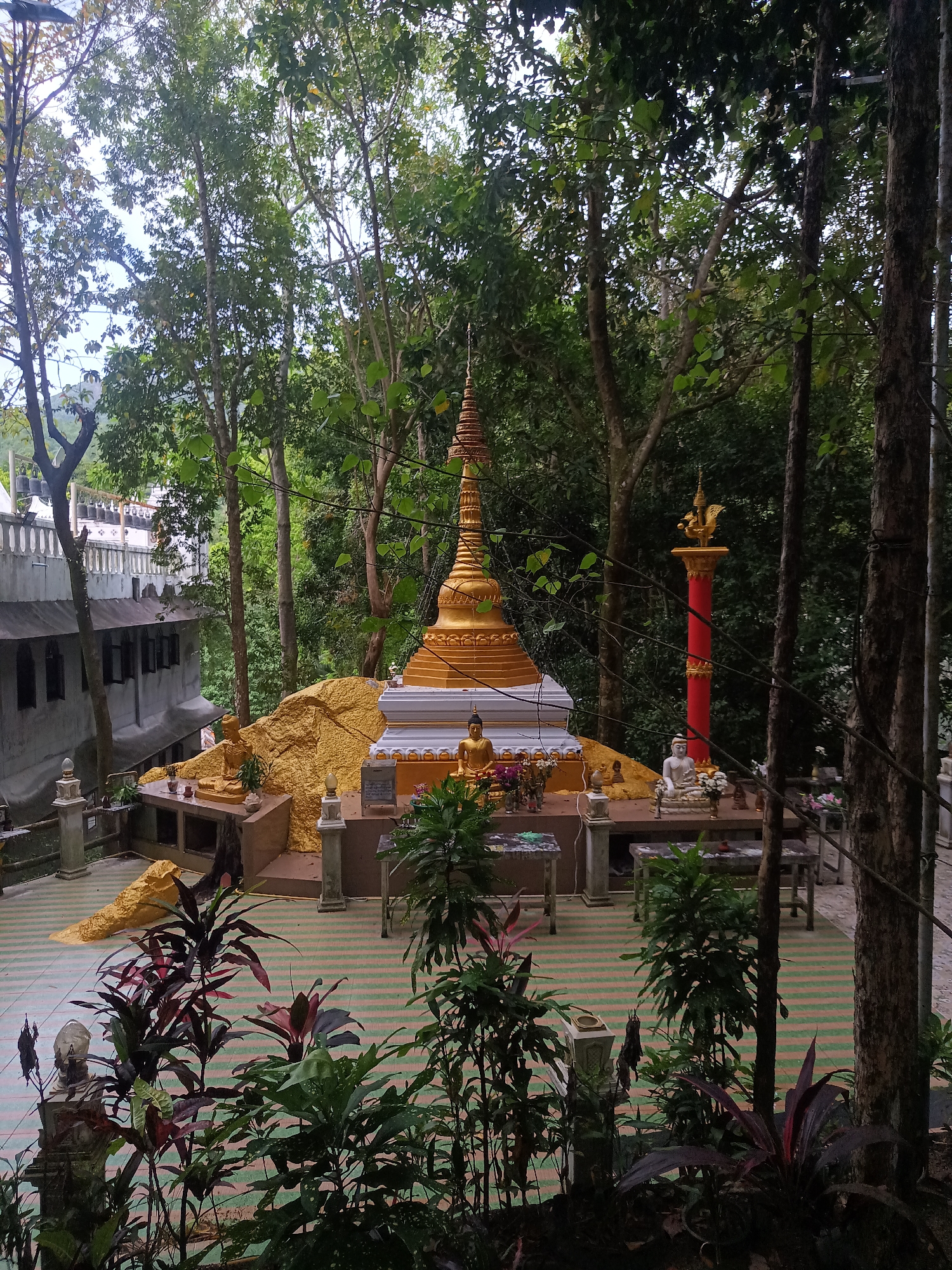 The image depicts a Buddhist shrine in a wooded area in ตำบลเกาะพะงัน, Thailand. A golden stupa stands centrally on a raised platform with golden statues around it. The platform has a tiled floor. A red and gold pillar stands to the right of the stupa. The background consists of lush trees and greenery. A building is visible on the left side. Tropical plants are located in the foreground.