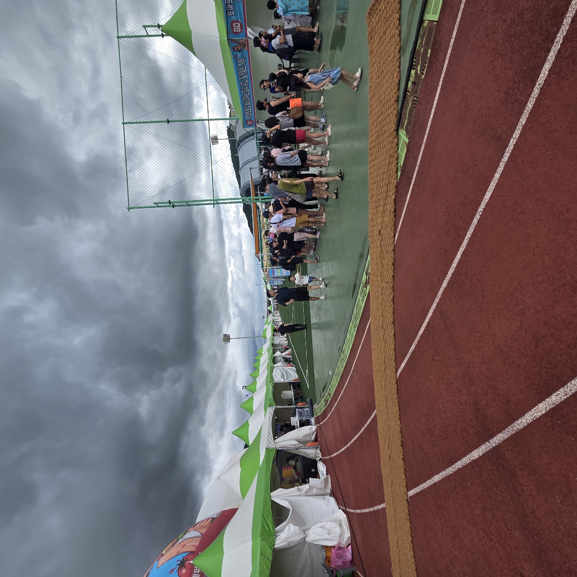 In Dunnae-myeon, South Korea, under a heavily overcast sky, a temporary outdoor market or festival is set up alongside a red running track. A line of white and green tents stretches into the distance, some with their fabric sides rolled up to reveal activity within. People of various ages are present, some browsing at the tents, others standing in groups, and a few are walking. In the foreground, a textured brown mat lies across the running track. The overall scene suggests a public event taking place outdoors, possibly before or during inclement weather.